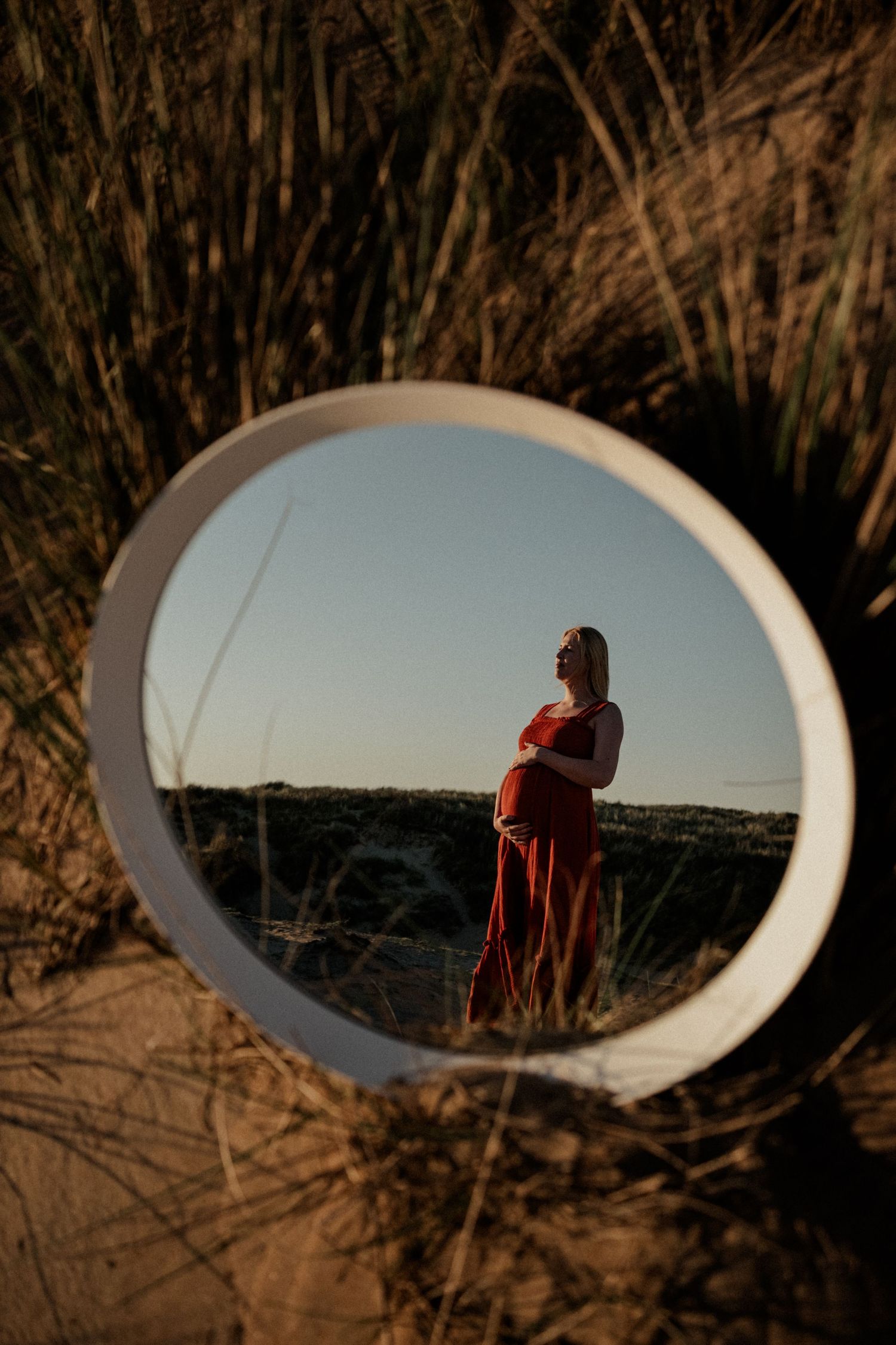 Pregnant woman in red dress reflected in round mirror among sand dunes at golden hour.