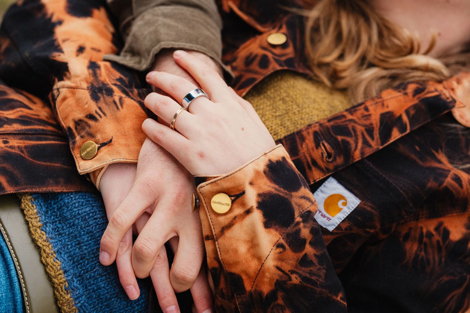 Close-up of two hands intertwined, one wearing a ring, over orange tie-dye Carhartt jackets.