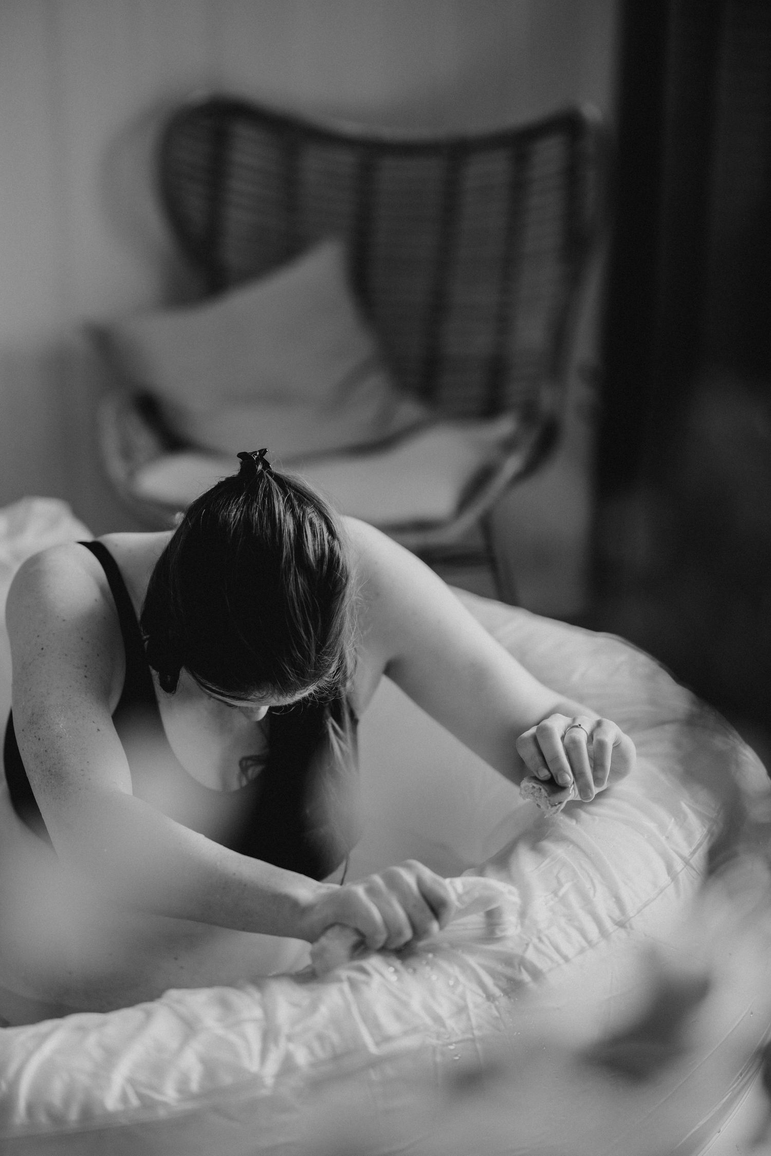 Black and white photo of a woman in lingerie lying on a bed, stretching forward with hands resting on white bedding.