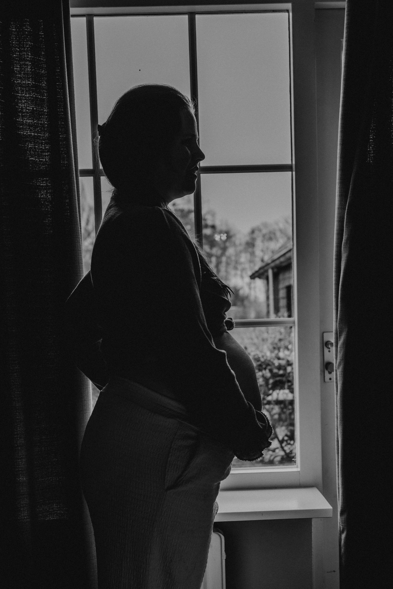 Black and white silhouette of a pregnant woman standing by a window, cradling her baby bump in natural light.