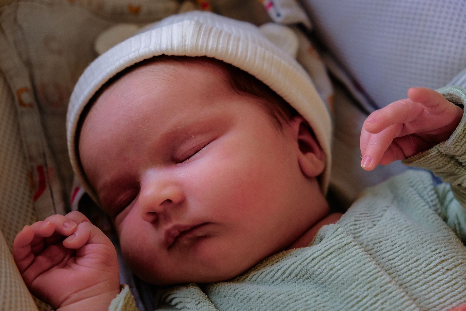 Newborn baby sleeping peacefully in hospital crib wearing white knit hat, with tiny hands visible.