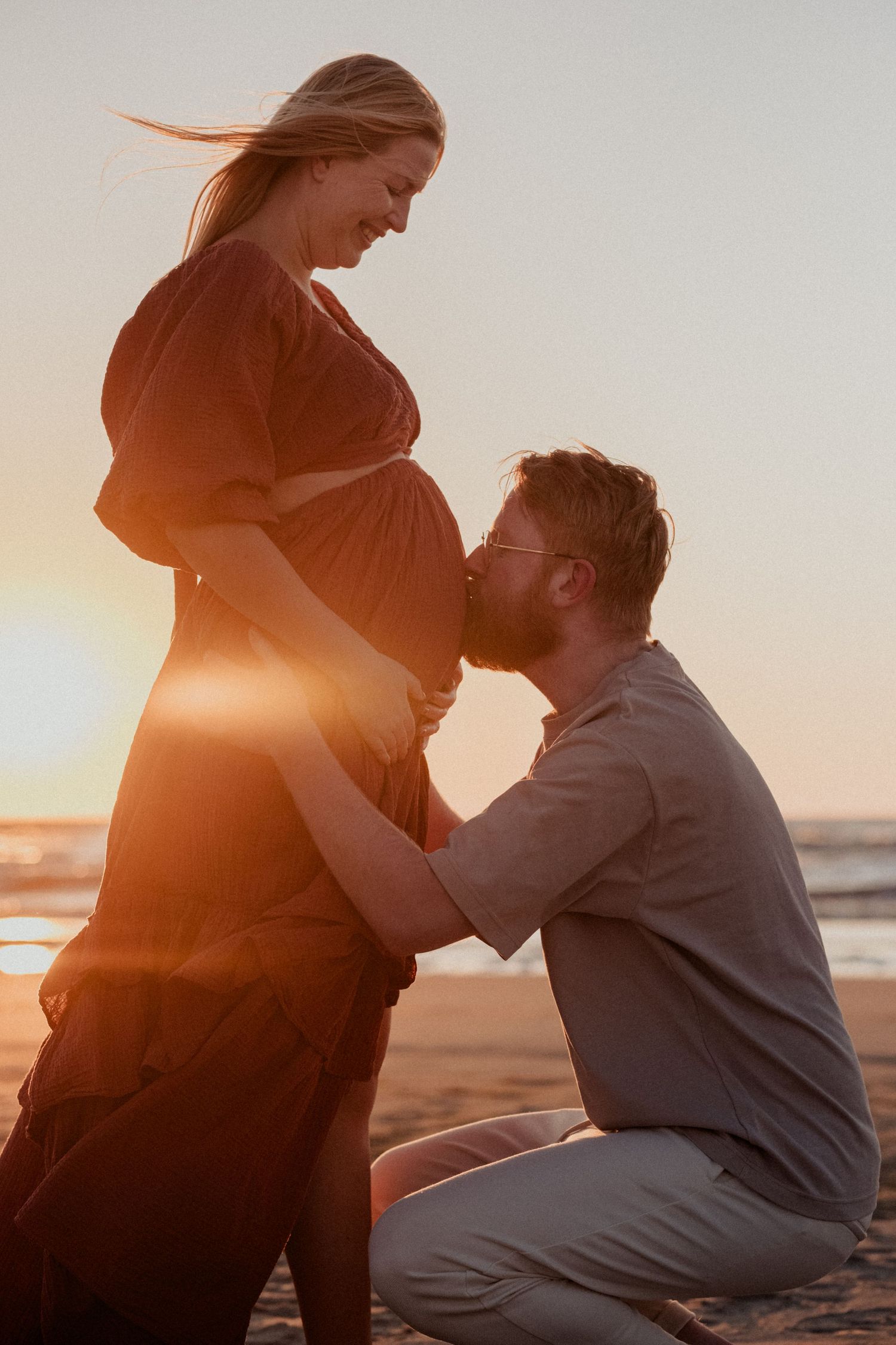 Man kneeling on beach kissing pregnant woman's belly at golden sunset, romantic maternity photo.