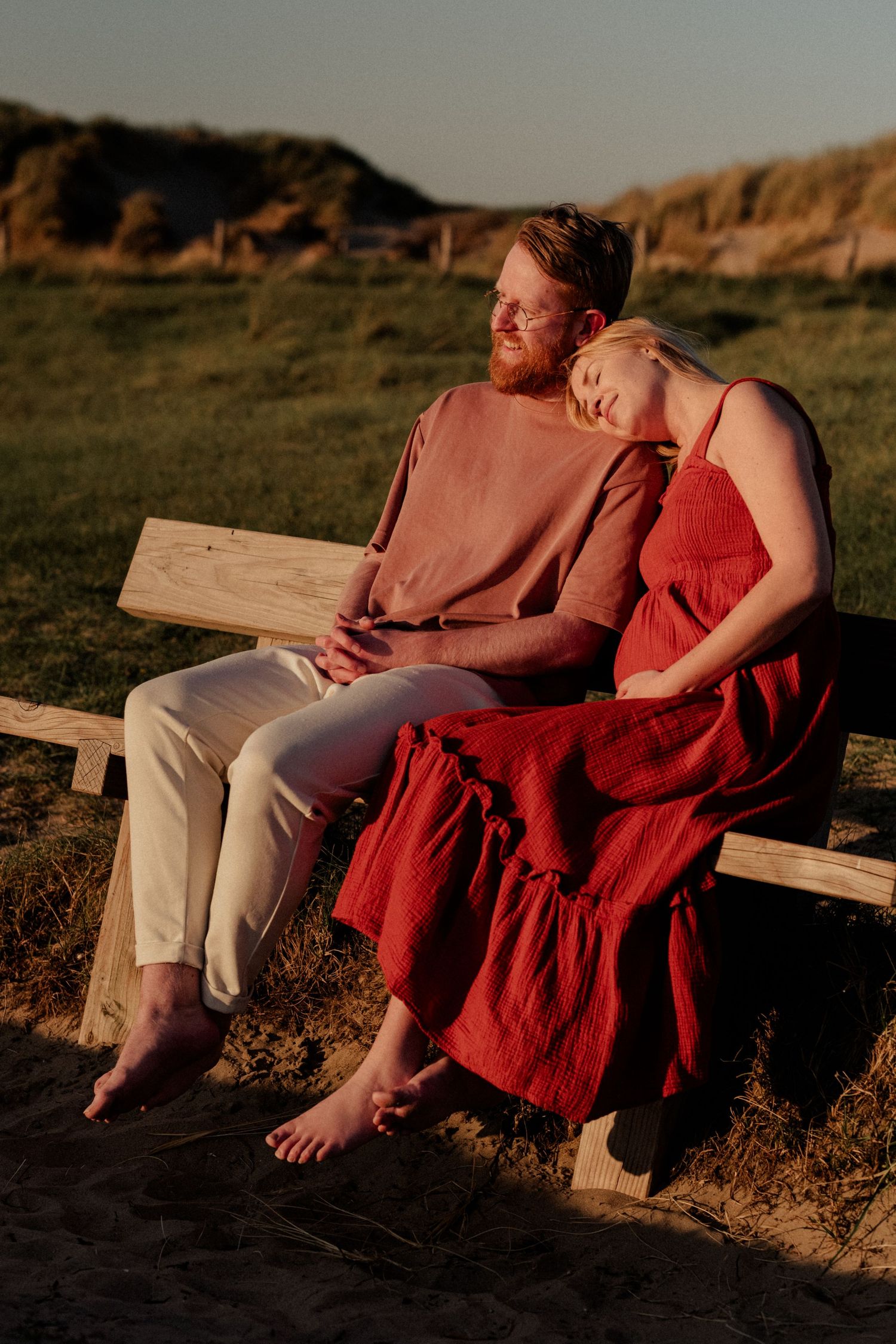 Couple sitting on wooden bench at sunset, woman in red dress resting head on man's shoulder outdoors.