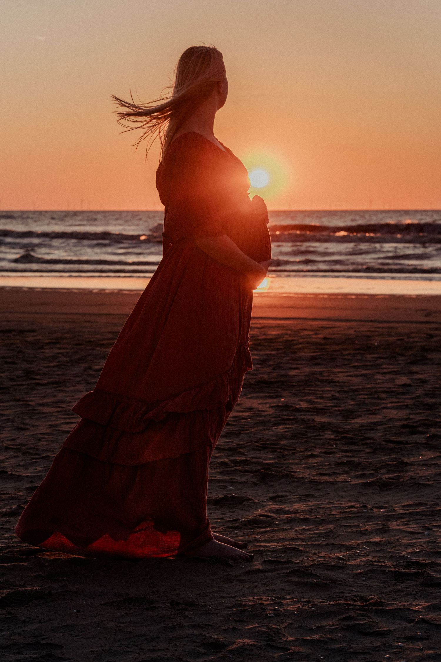 Pregnant woman in red dress silhouetted against golden sunset on beach with flowing hair and ocean backdrop.