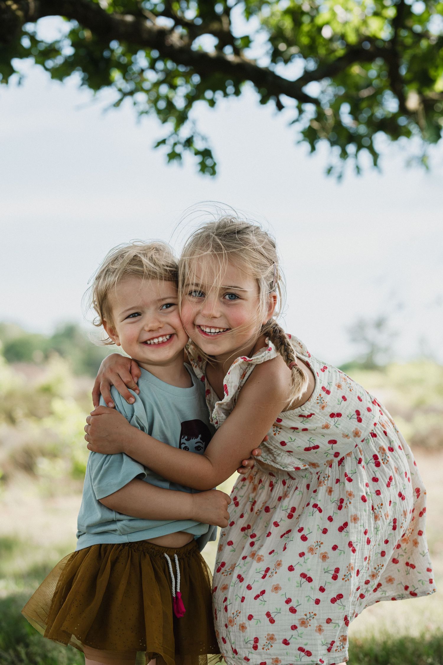 Two young sisters hugging and smiling outdoors under a tree on a sunny day.