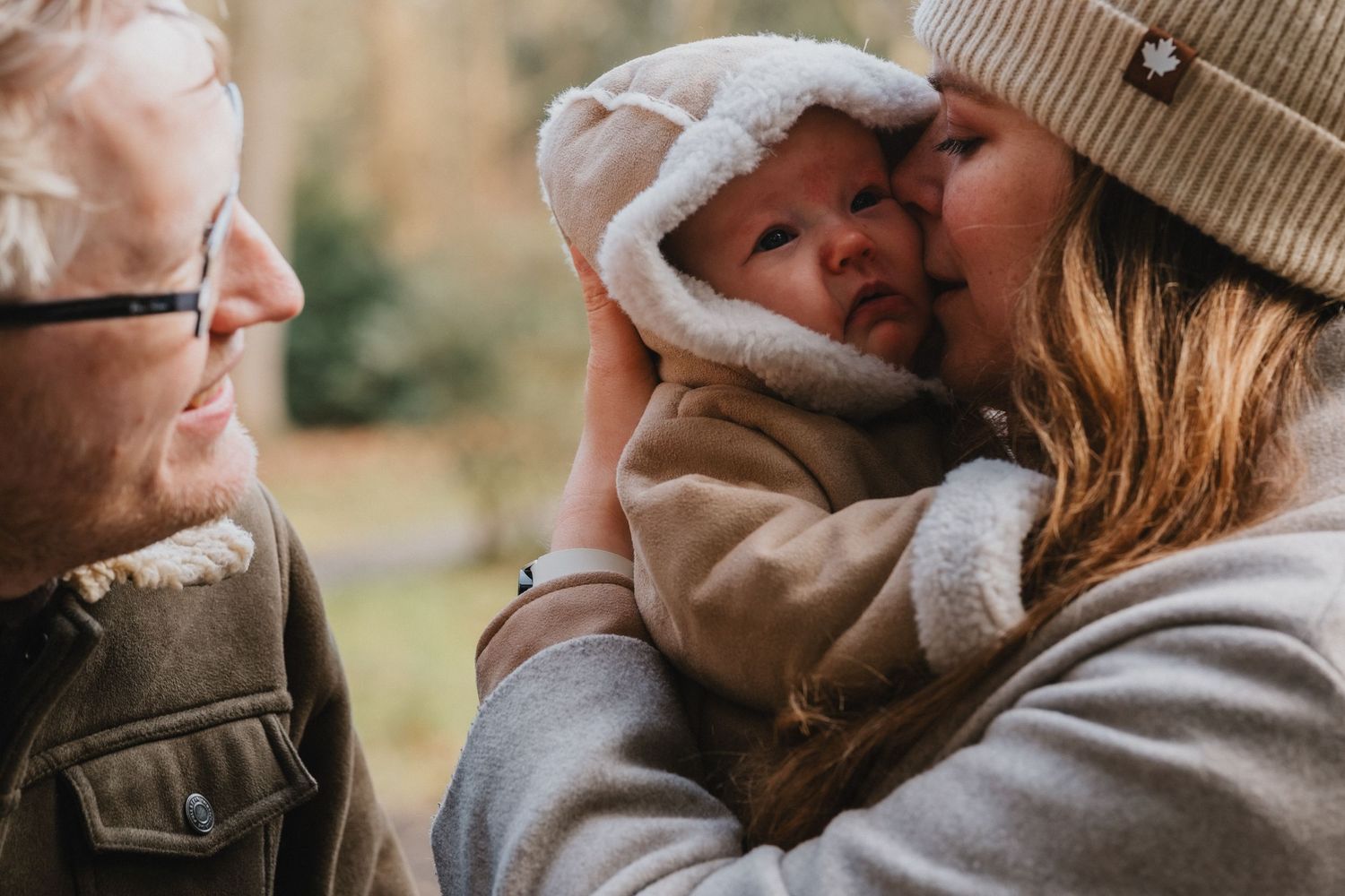 A couple in autumn outdoor setting tenderly holding their newborn baby wrapped in a cozy hooded jacket.