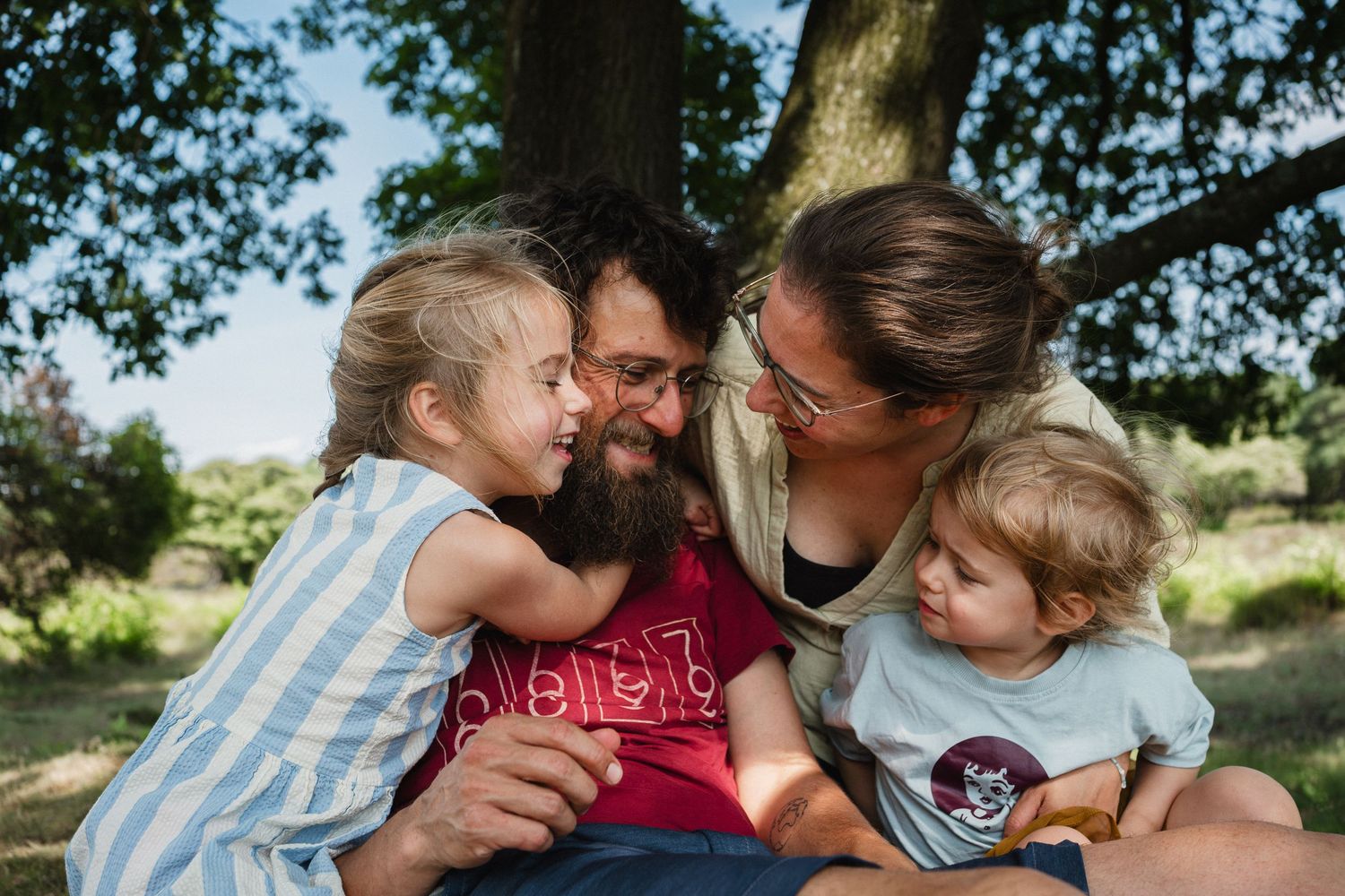 Happy family with two young children sharing a joyful moment outdoors under a tree on a sunny day.