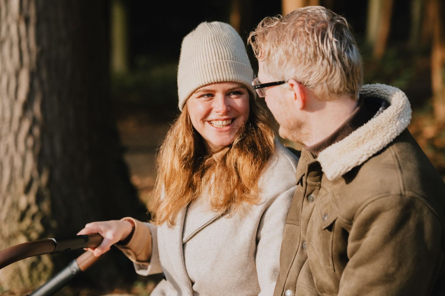 A smiling couple sitting together outdoors in a forest, woman in beige coat and knit hat, man in sherpa jacket.