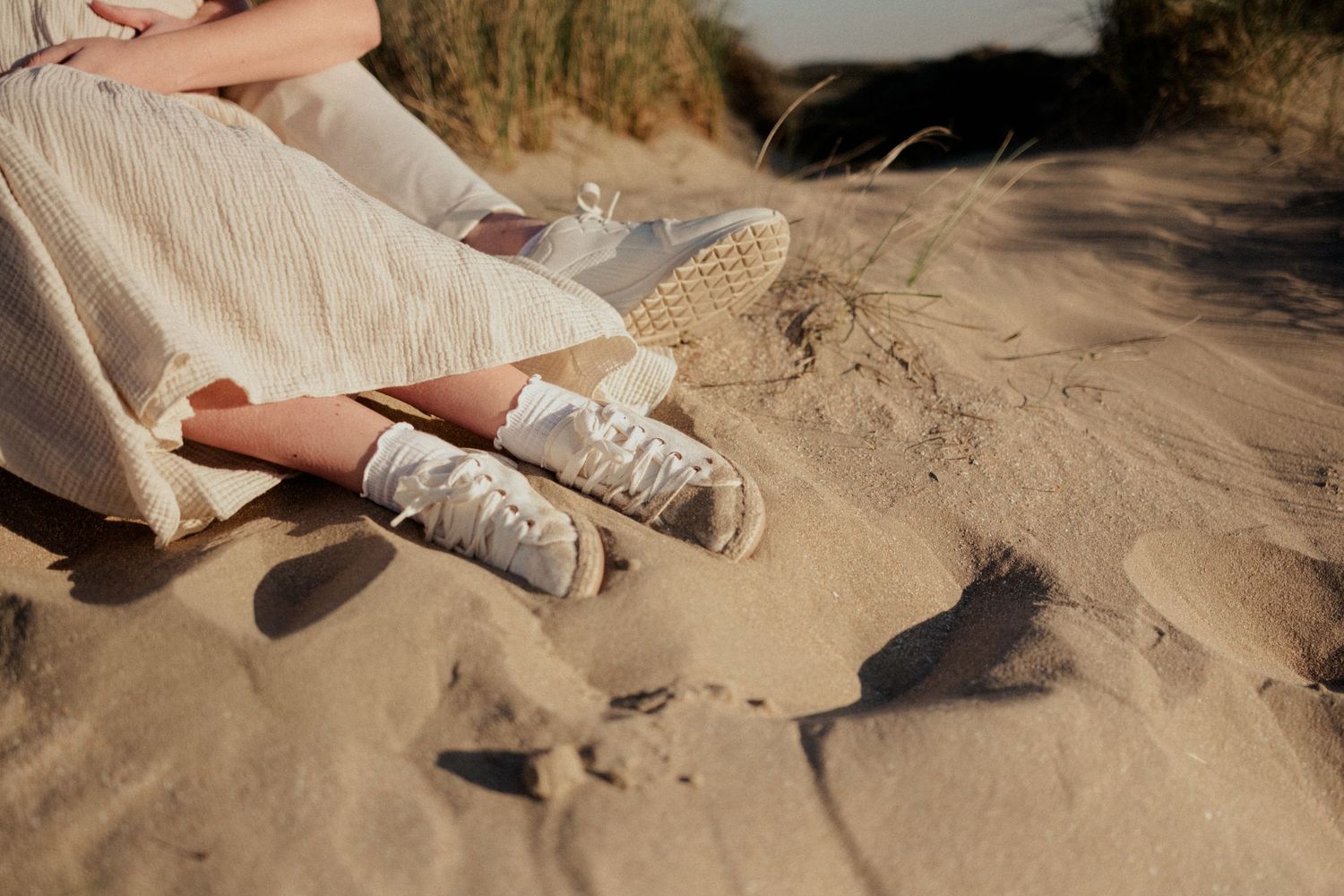Person sitting on sandy dunes wearing a white linen dress and lace socks with woven bag beside them.
