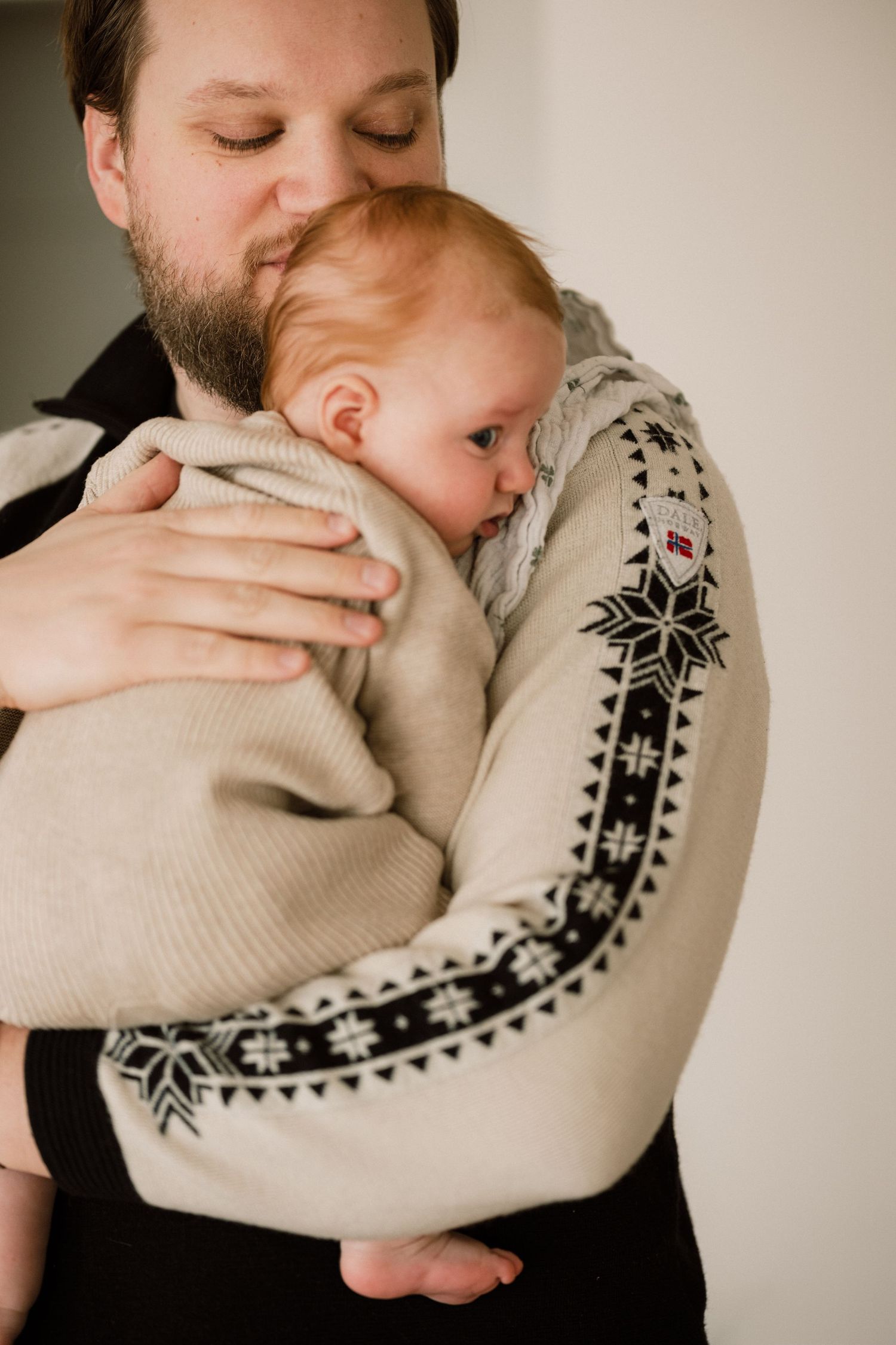 Bearded father in Nordic sweater tenderly holding and kissing his sleeping newborn baby.