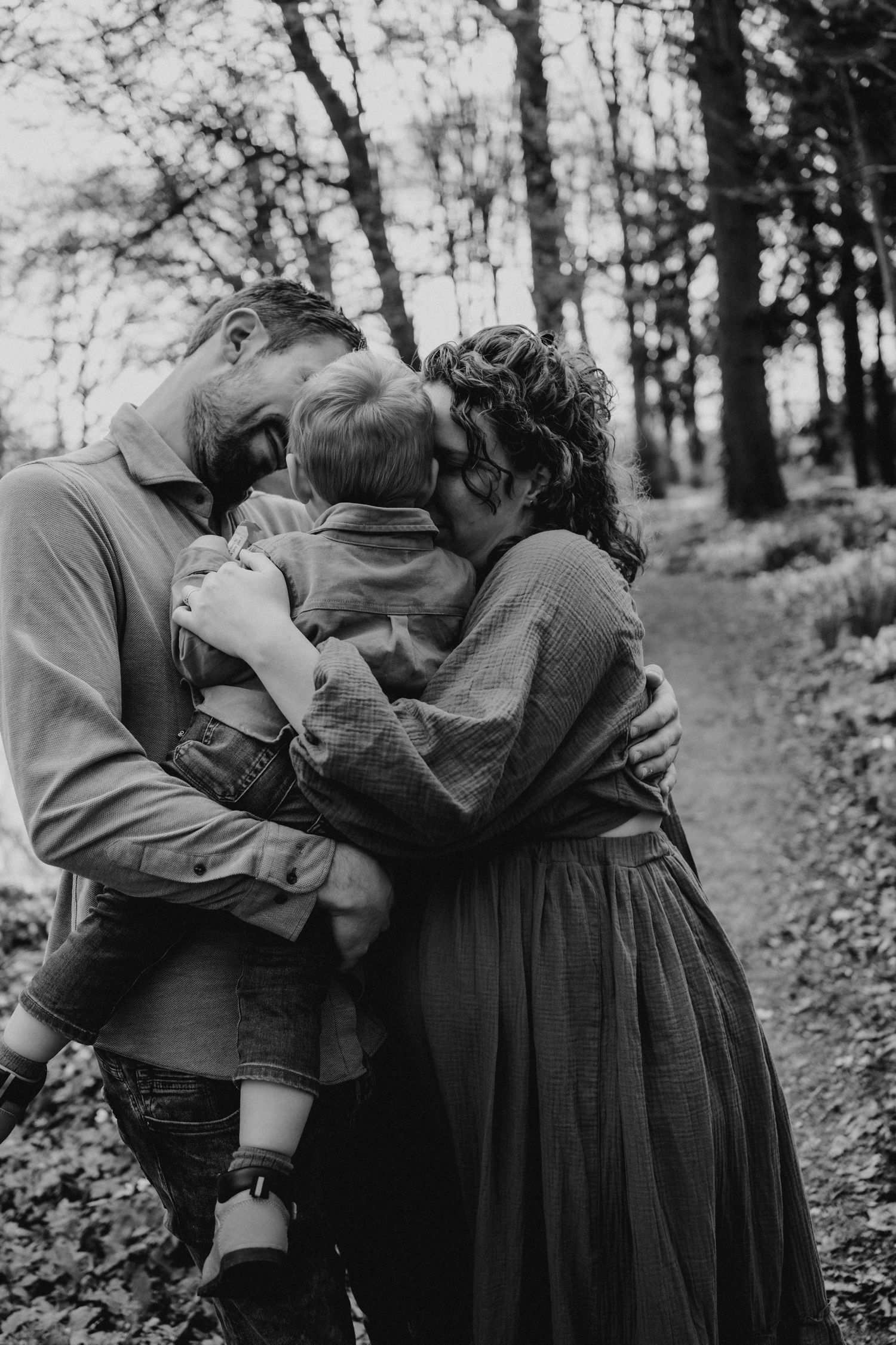 Black and white photo of parents lovingly embracing their toddler in a woodland path setting.