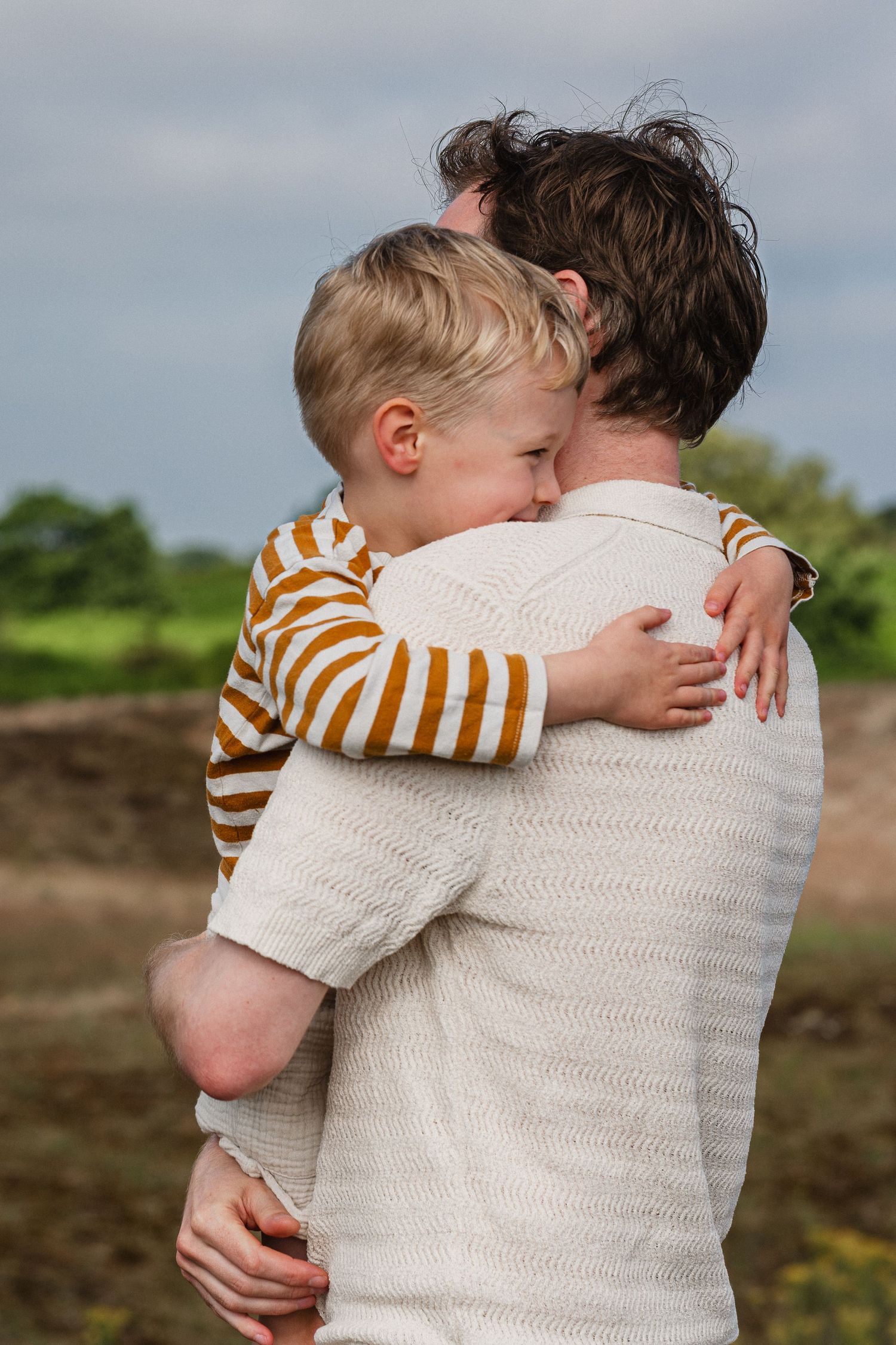 A young blonde child hugging an adult outdoors, wearing a striped orange shirt in a warm embrace.