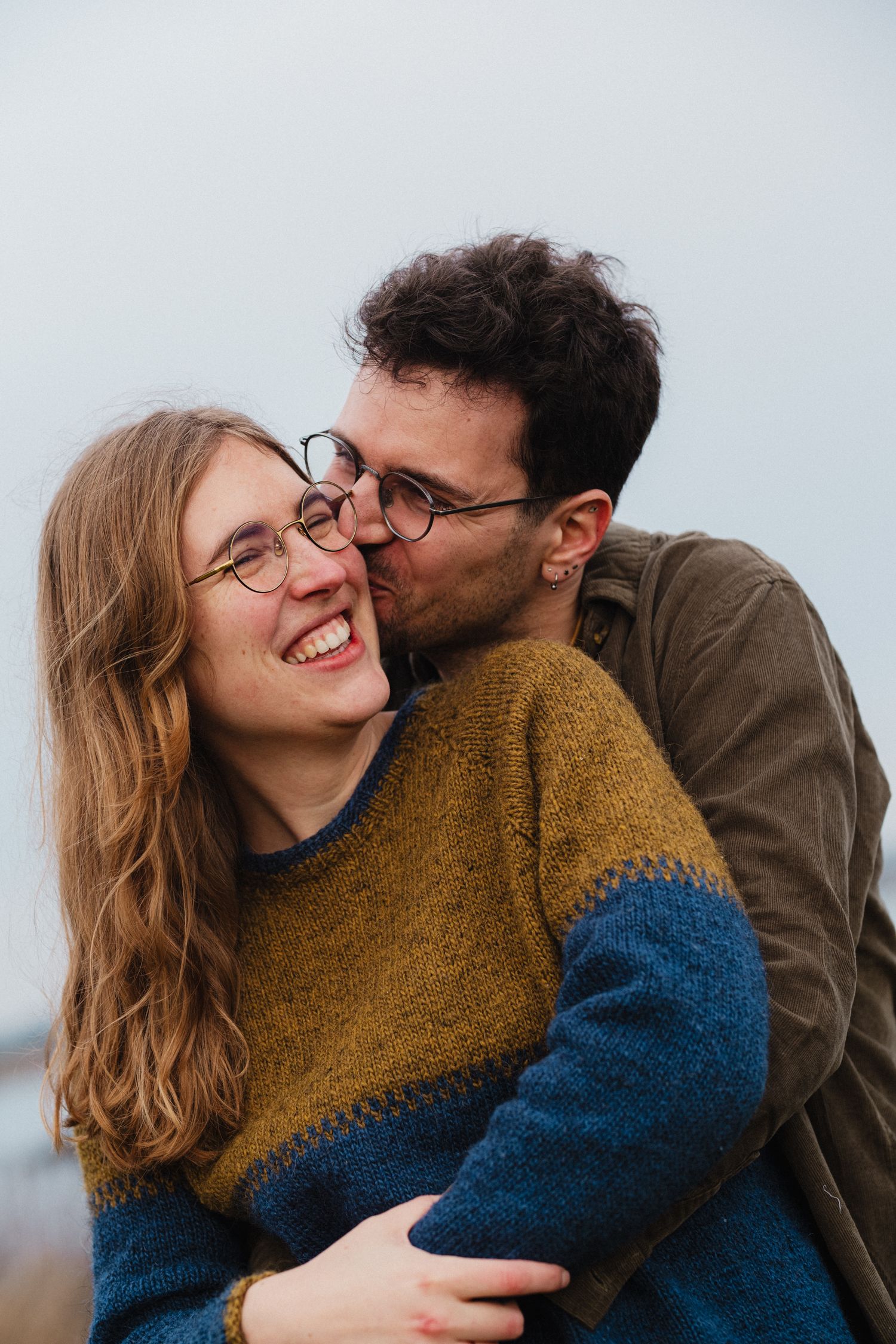 A smiling woman with glasses receives a kiss on the cheek from a man hugging her from behind outdoors.