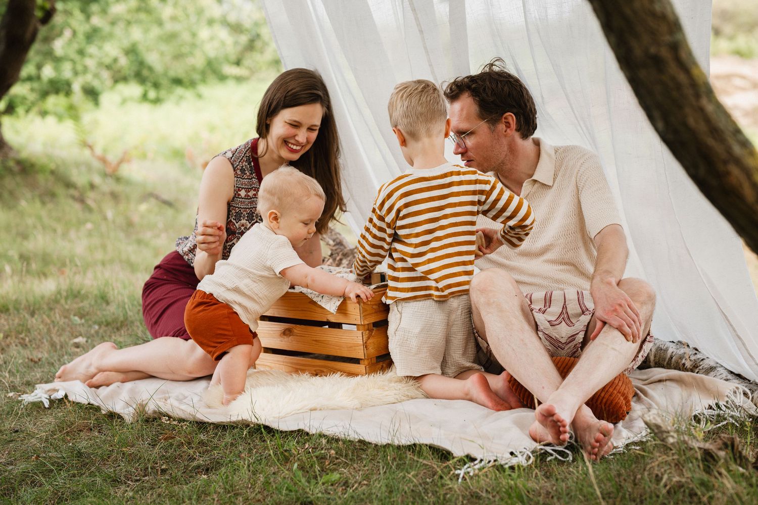 Family of four sitting on blanket outdoors near white teepee, parents with two young children playing with wooden crate.