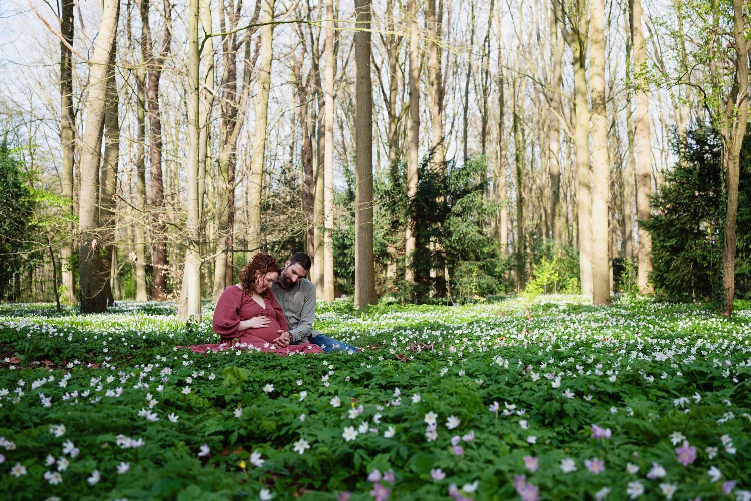 Couple sitting together in a blooming spring forest with white wildflowers and tall bare trees in a romantic outdoor setting.