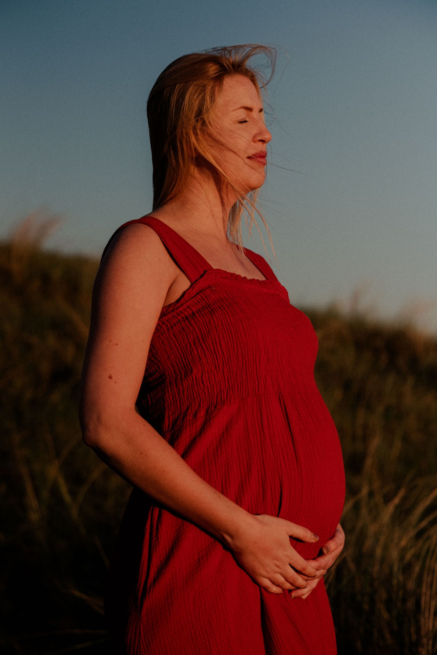 Pregnant woman in red dress cradling her baby bump outdoors at golden hour with wind in her blonde hair.