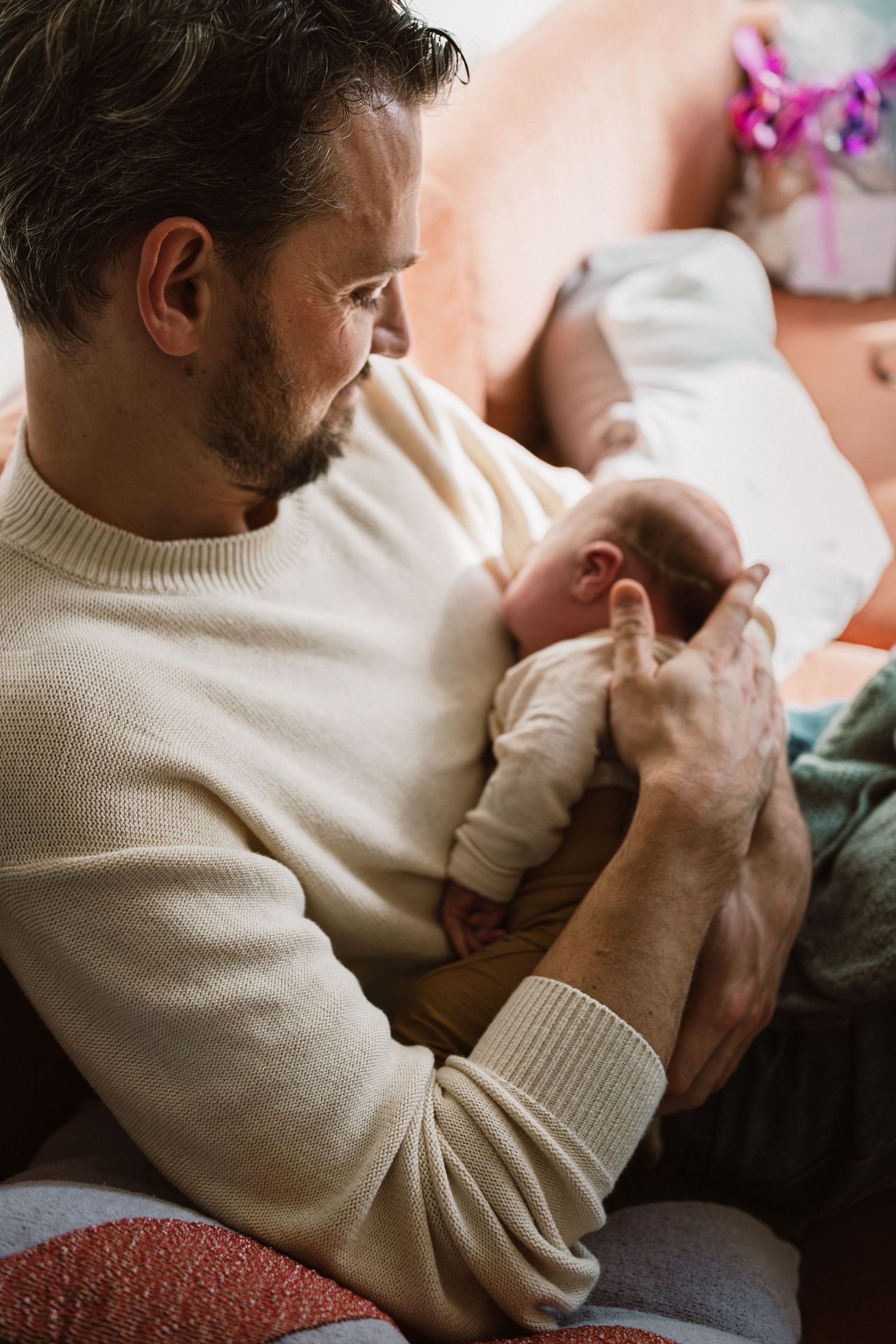 A bearded man in a cream sweater tenderly holds a newborn baby while sitting on a couch.