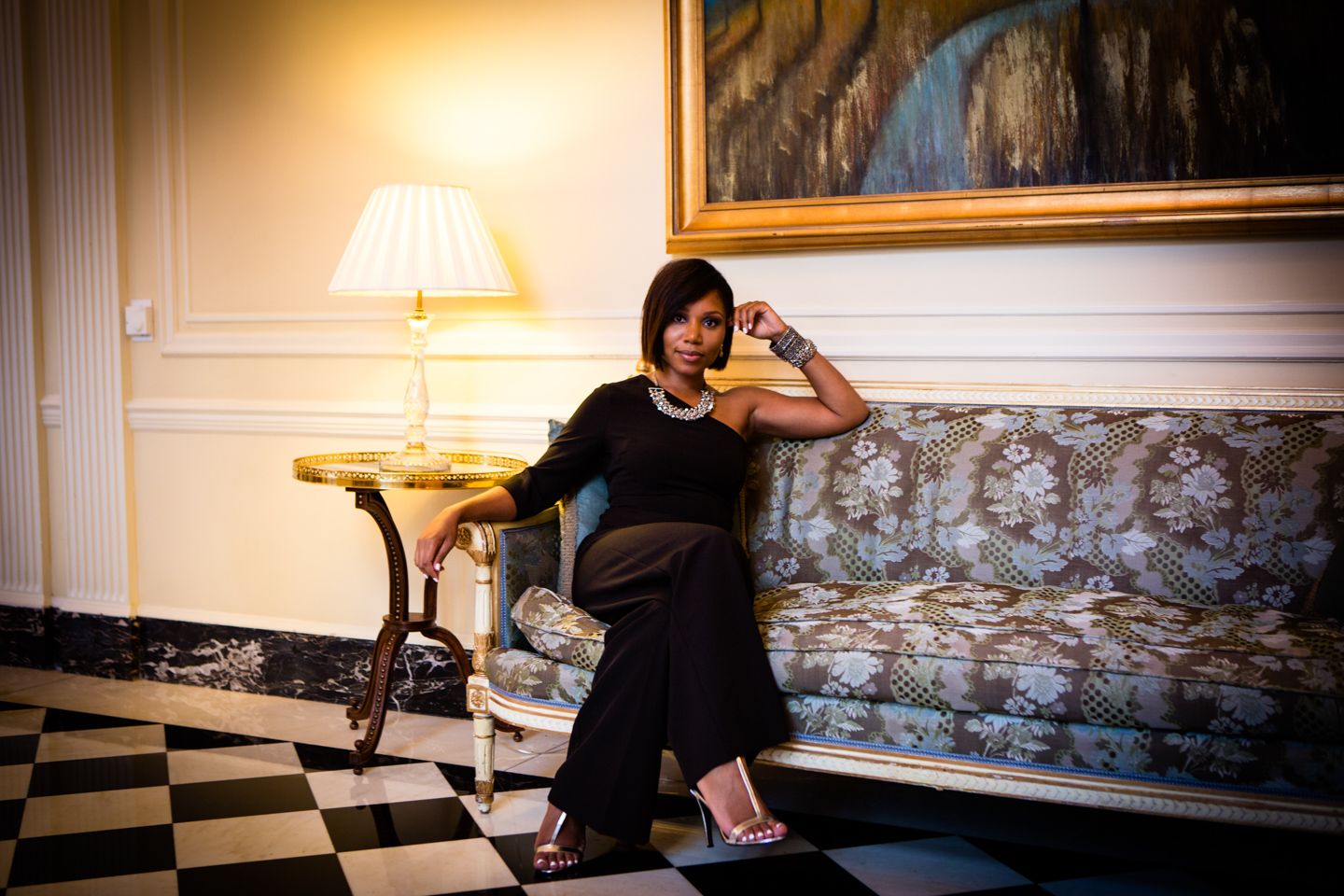 Black woman in a black dress relaxes on an antique sofa in an elegant room with checkerboard floors and warm lighting.