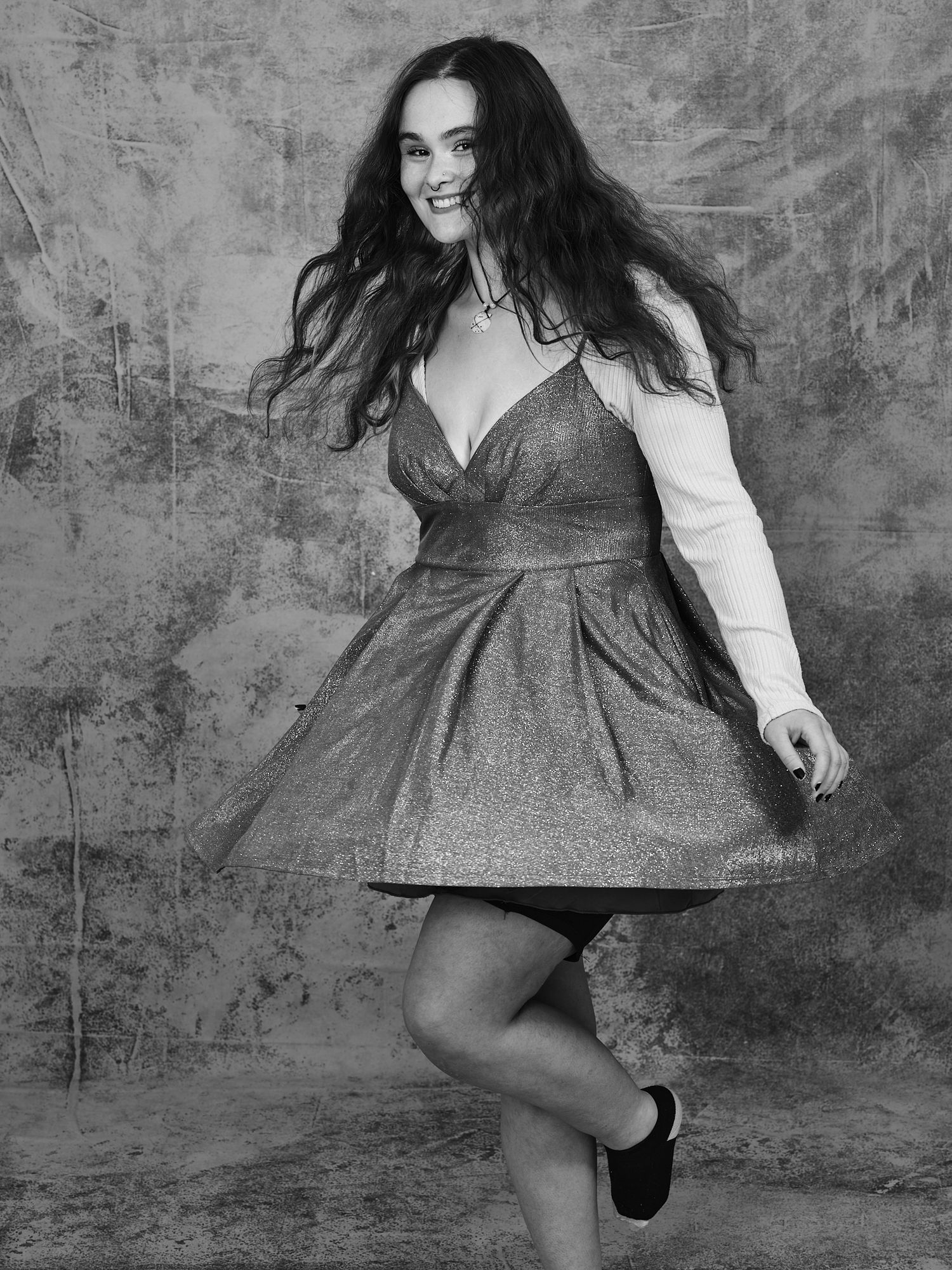 Joyful dancing pose in a sparkly dress and white long-sleeve top against textured background.