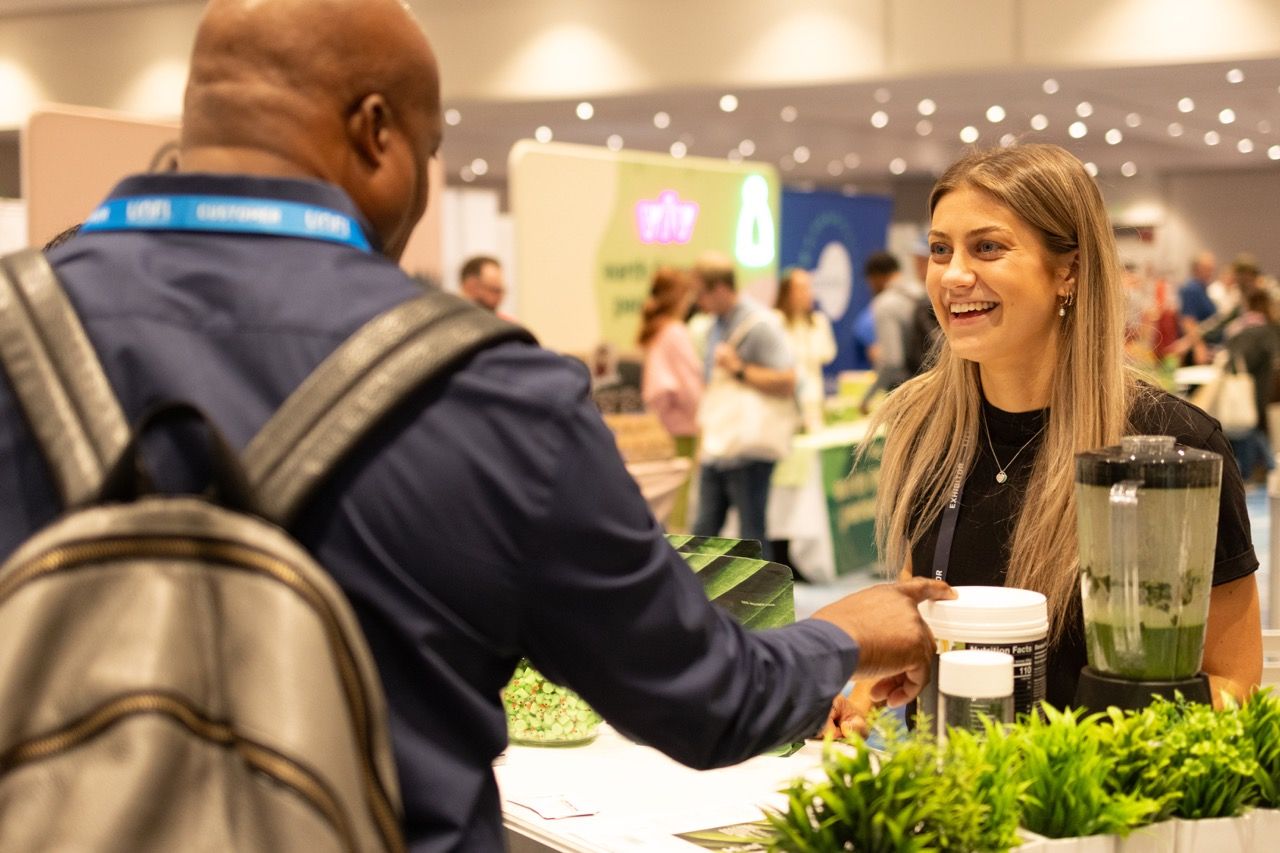 A woman smiles at a trade show booth featuring green plants and a blender, engaging with a visitor.