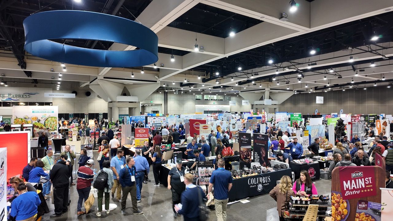 Busy trade show expo floor filled with vendors, booths, and attendees browsing products in a large convention center.