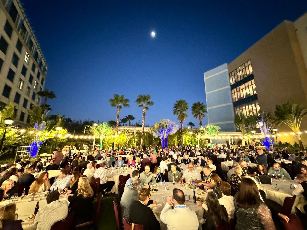 Elegant outdoor evening gala with guests dining under palm trees and a moonlit sky near a modern building.