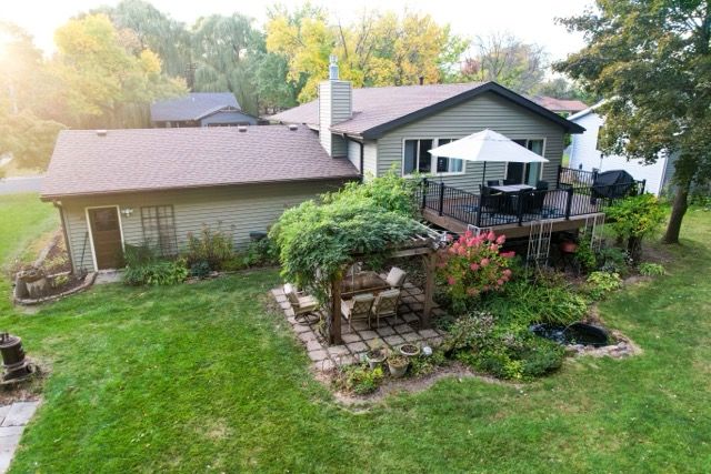 Aerial view of a suburban home with a wooden deck, patio umbrella, lush garden beds, and green lawn surrounded by trees.