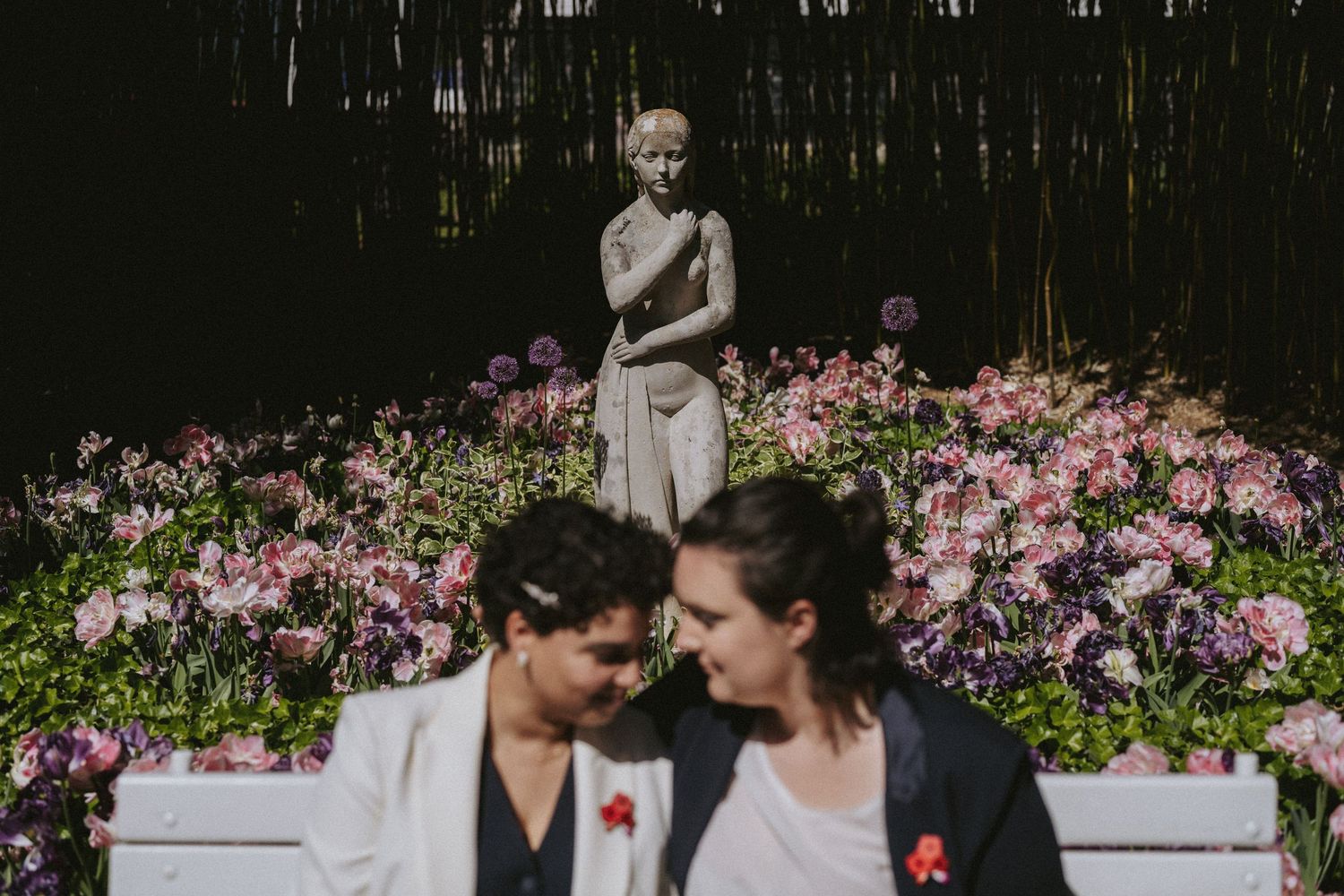 Two brides share a tender moment on a white bench surrounded by pink flowers and a garden statue.