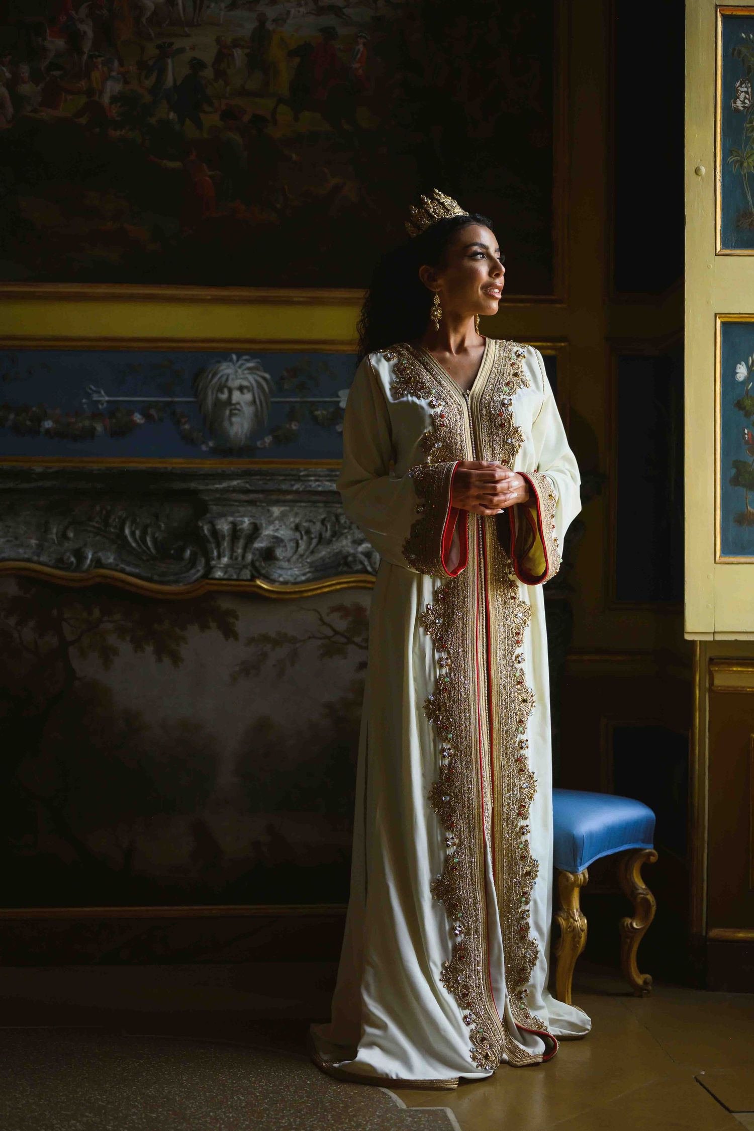 Woman in elegant white embroidered kaftan gown with gold crown standing in ornate room with classical paintings.