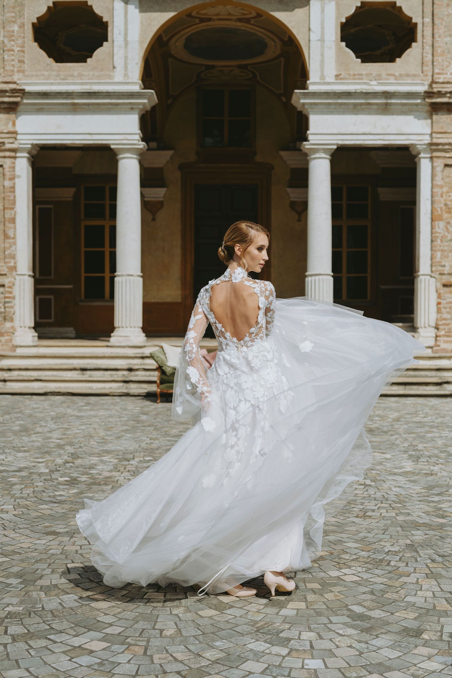 Bride in a flowing white lace wedding gown with open back twirling in a cobblestone courtyard of an elegant venue.