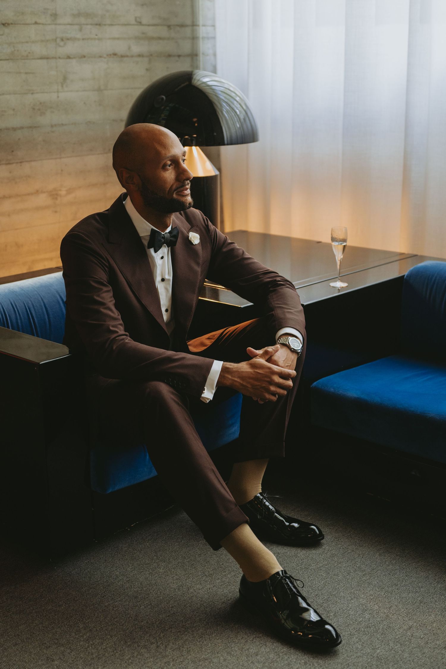 Elegant bald man in dark burgundy tuxedo with bow tie sits on blue velvet sofa in stylish modern interior.