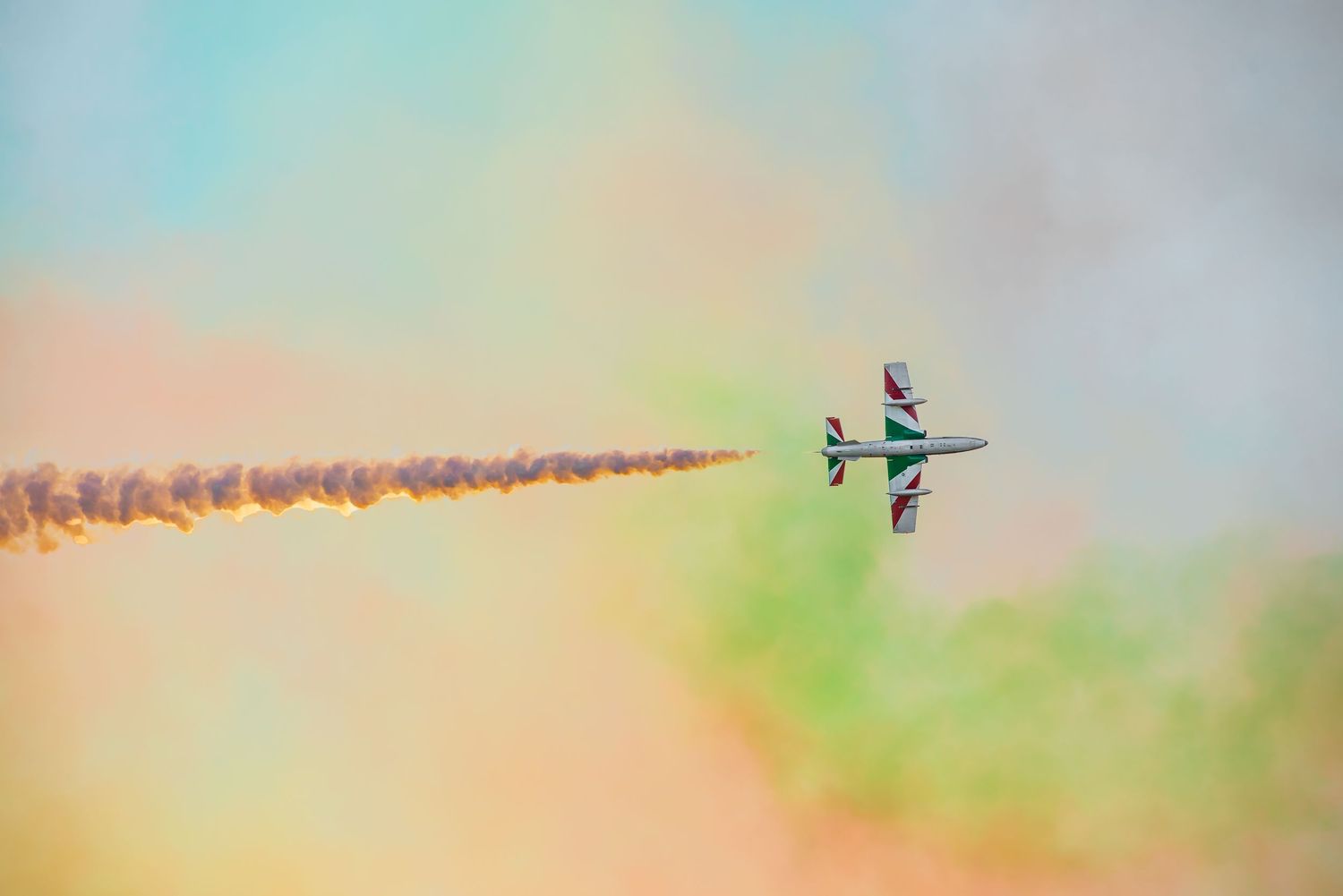 Colorful aerobatic jet aircraft performing at an airshow, trailing vibrant orange and green smoke against a pastel sky.