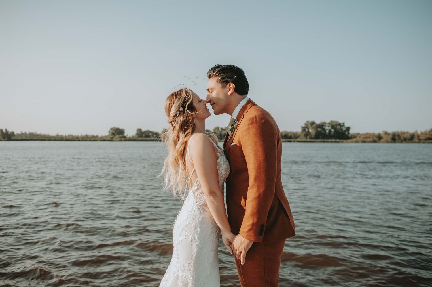 Bride and groom holding hands by a lake, wearing white lace gown and rust suit, sharing a romantic moment outdoors.