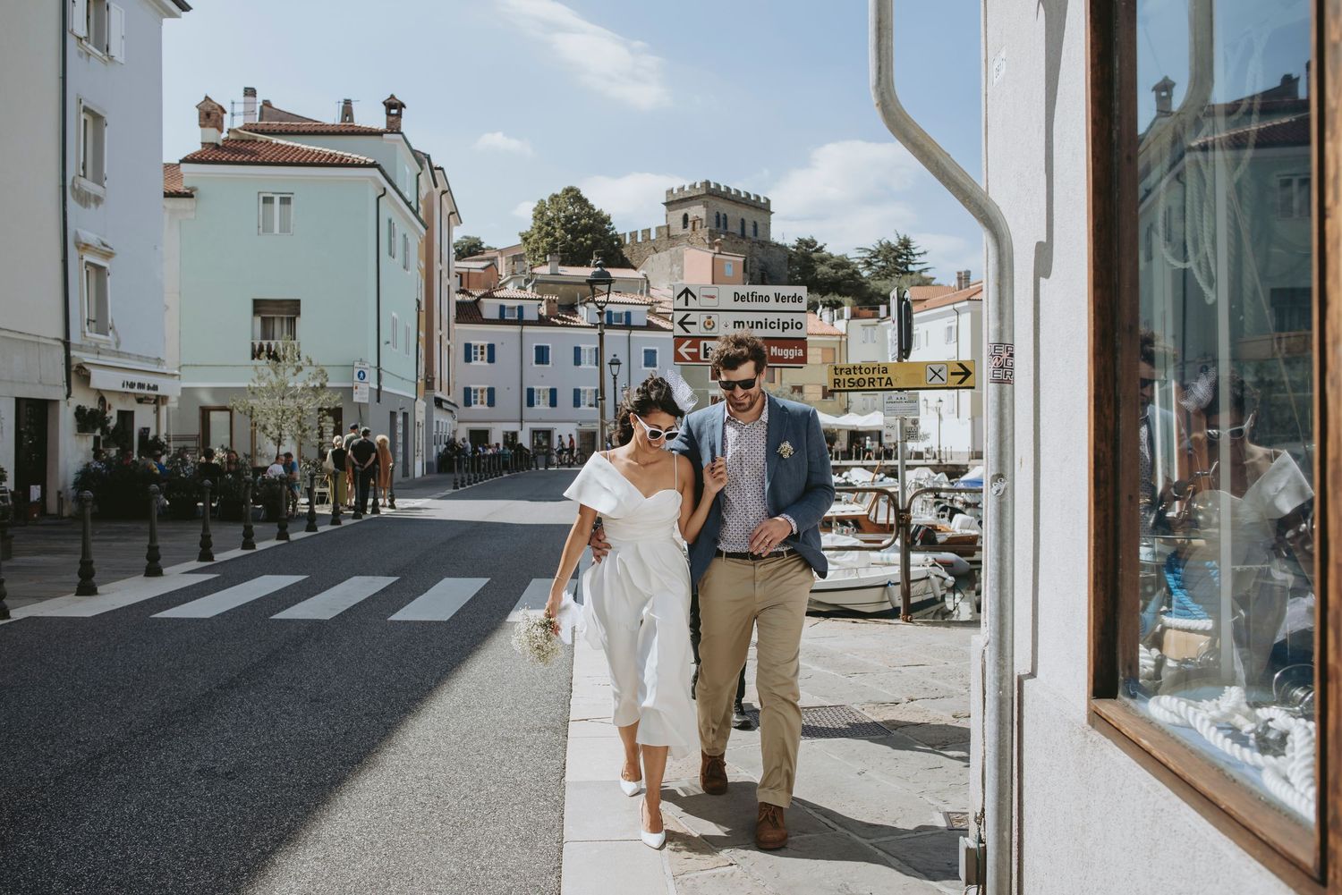 People walking on a sunny street with white buildings and storefronts in a Mediterranean coastal town.