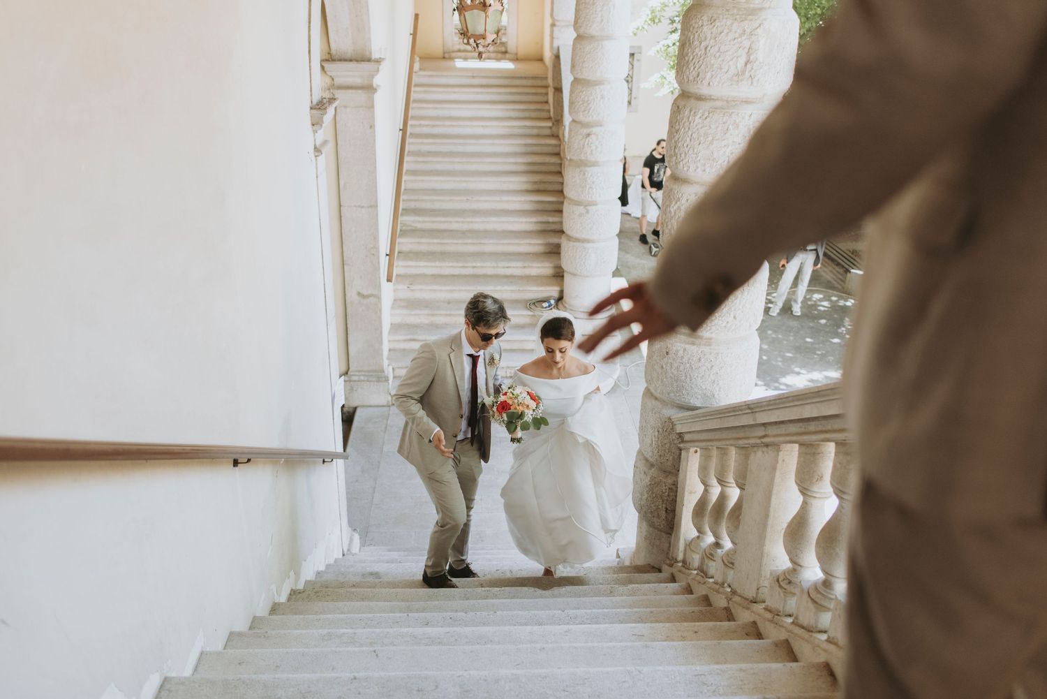 A couple in wedding attire ascends an elegant white marble staircase in a historic building.