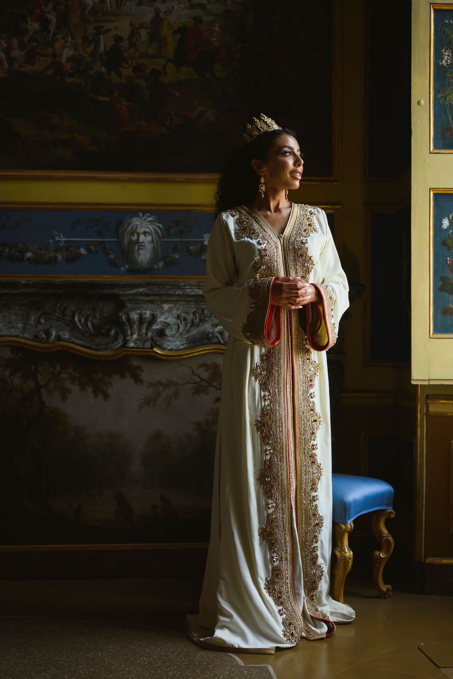 A person in an elegant cream-colored wedding dress with lace details stands in a dimly lit vintage room.