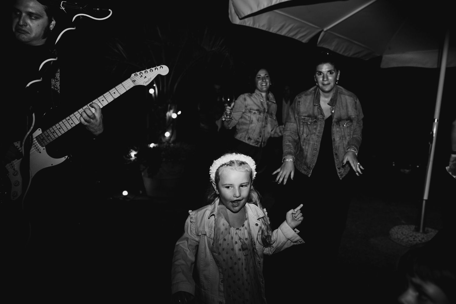 A black and white image showing a group of people, including a person playing an electric guitar, in a dimly lit setting.