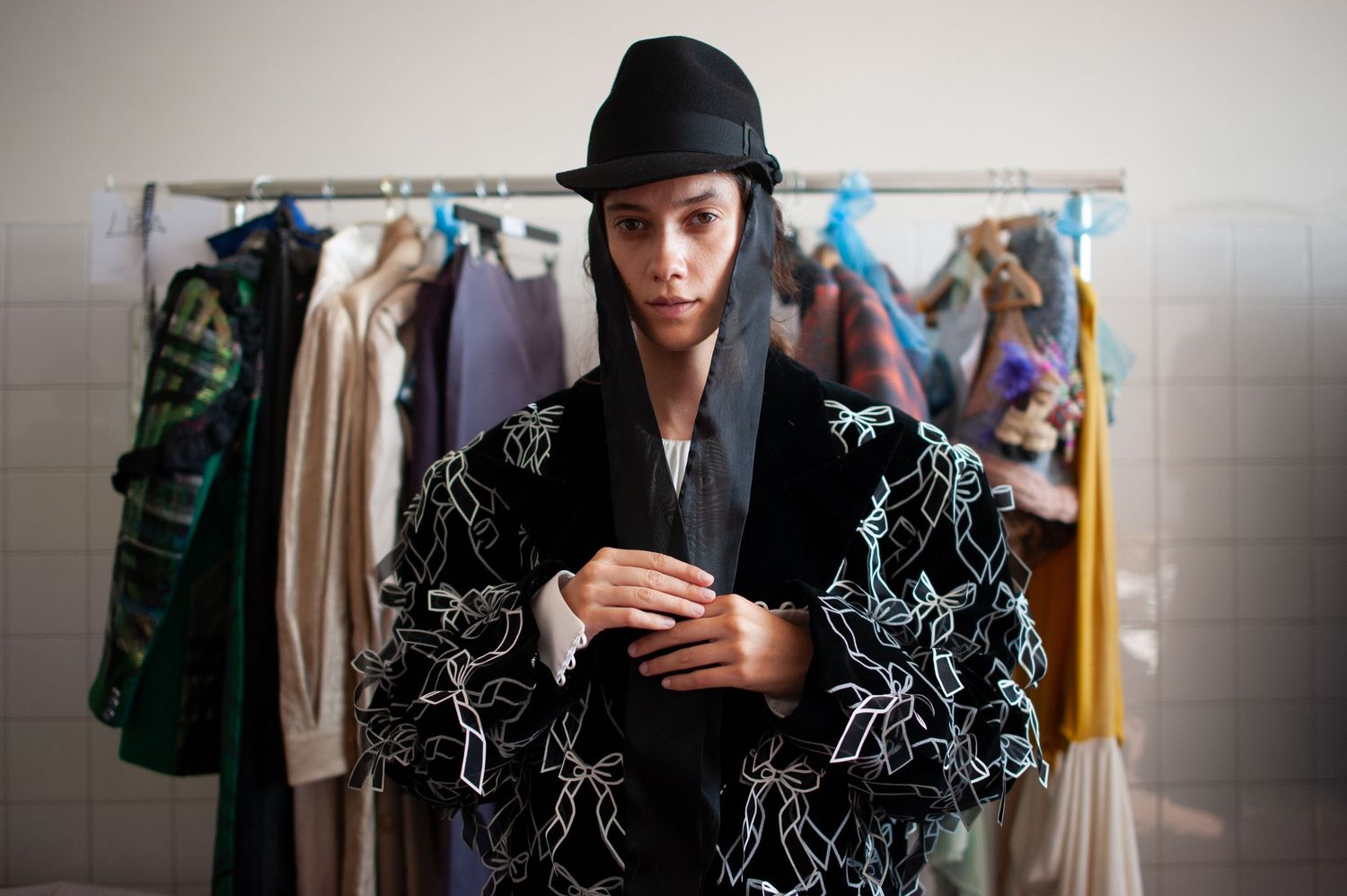 A model wearing a black embroidered jacket and hat stands before a clothing rack in a fashion backstage setting.