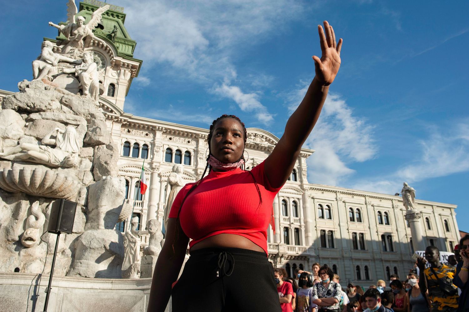 Woman in red top raises hand at outdoor protest near ornate European fountain and building.