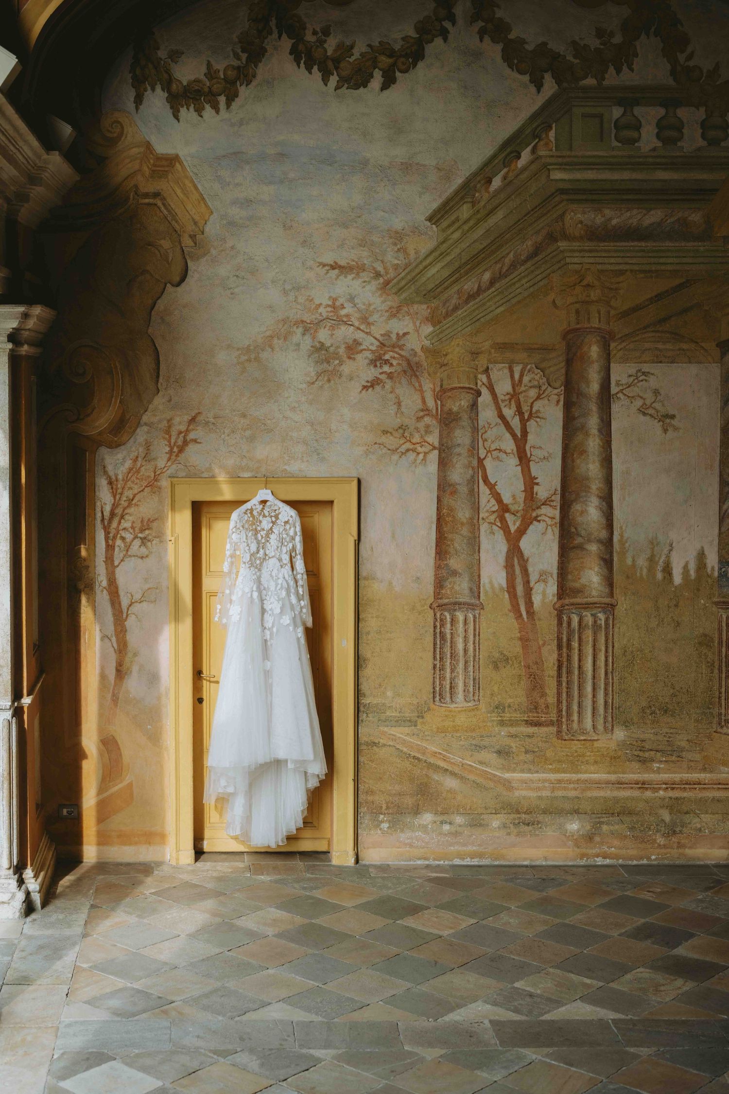 A white lace wedding dress hanging on a golden door frame against an ornate fresco-painted wall in a historic villa.