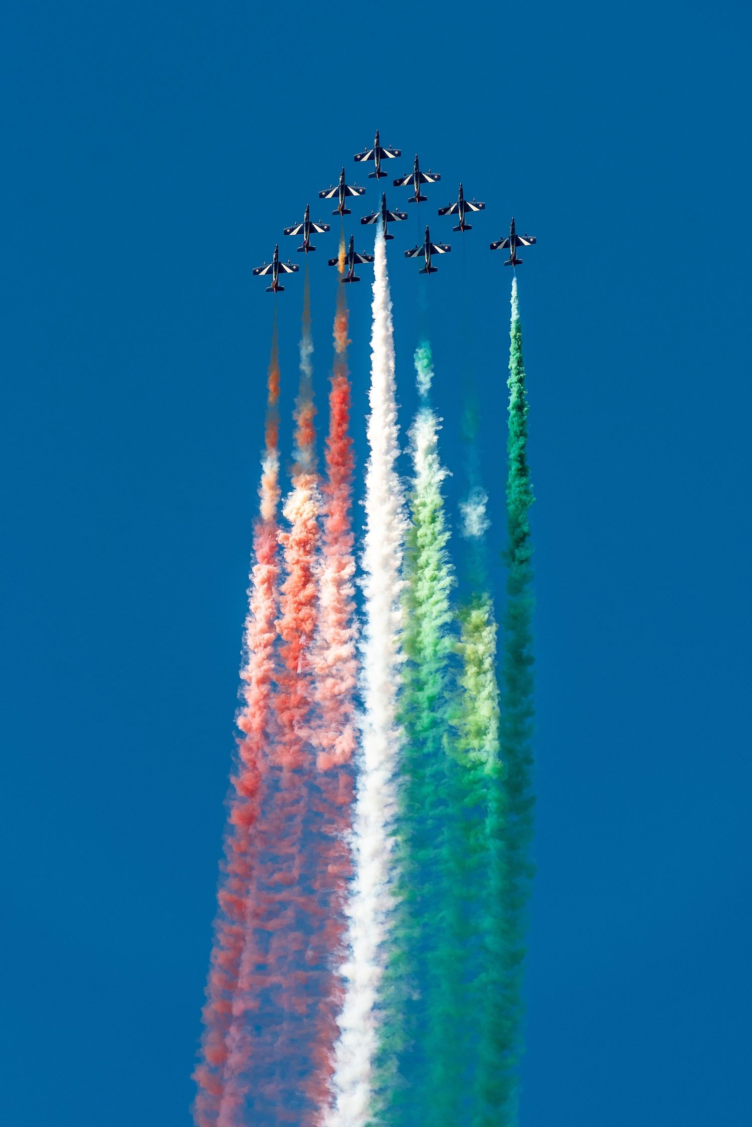 Italian aerobatic team Frecce Tricolori jets fly in formation releasing red, white, and green smoke trails against blue sky.
