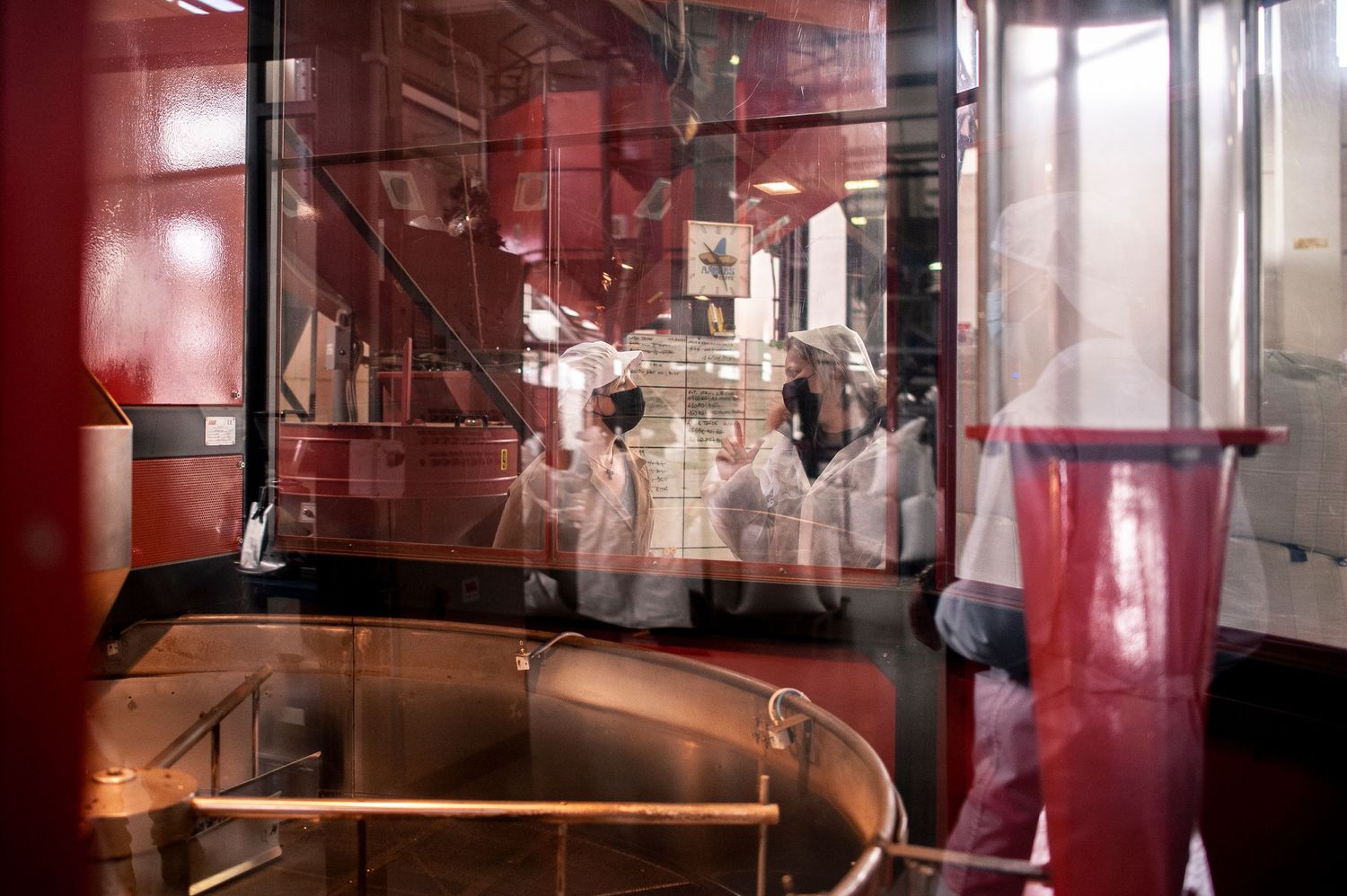 Workers in white coats inside a modern industrial brewery facility with red metallic equipment and glass panels.