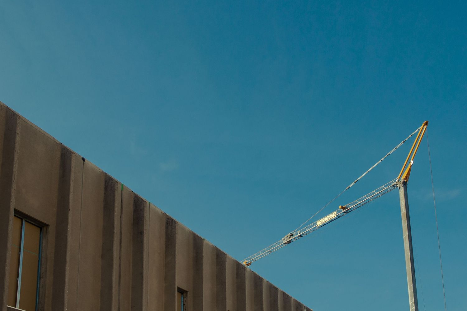 A construction crane extends over a modern building facade against a clear blue sky.