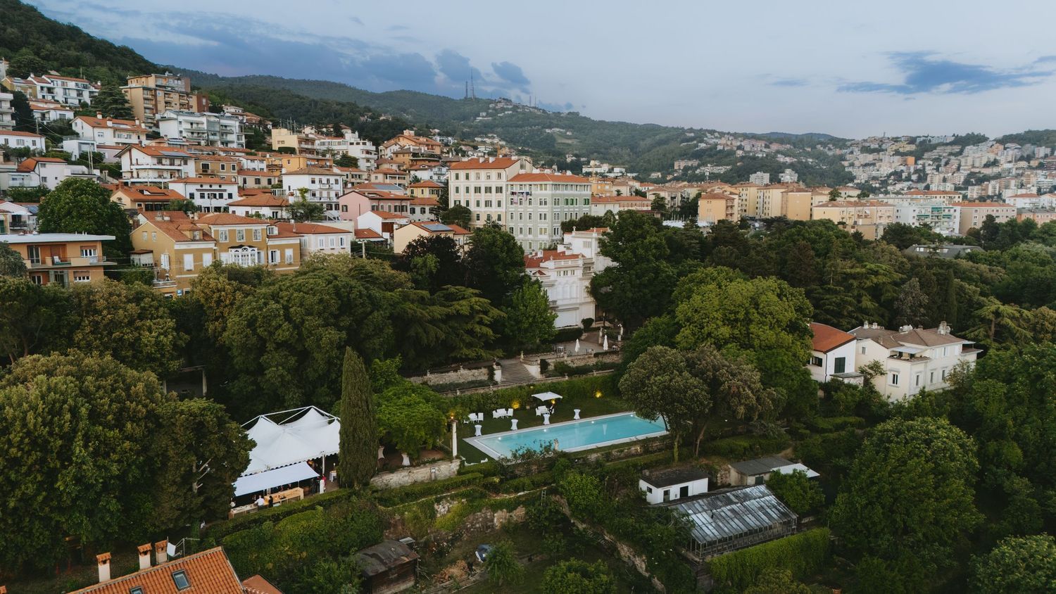 Aerial view of a Mediterranean hillside town with lush greenery, white buildings, and a pool surrounded by event tents.