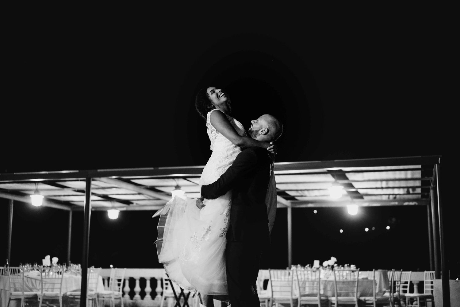 Groom lifts bride during first dance at outdoor wedding reception in black and white photography.