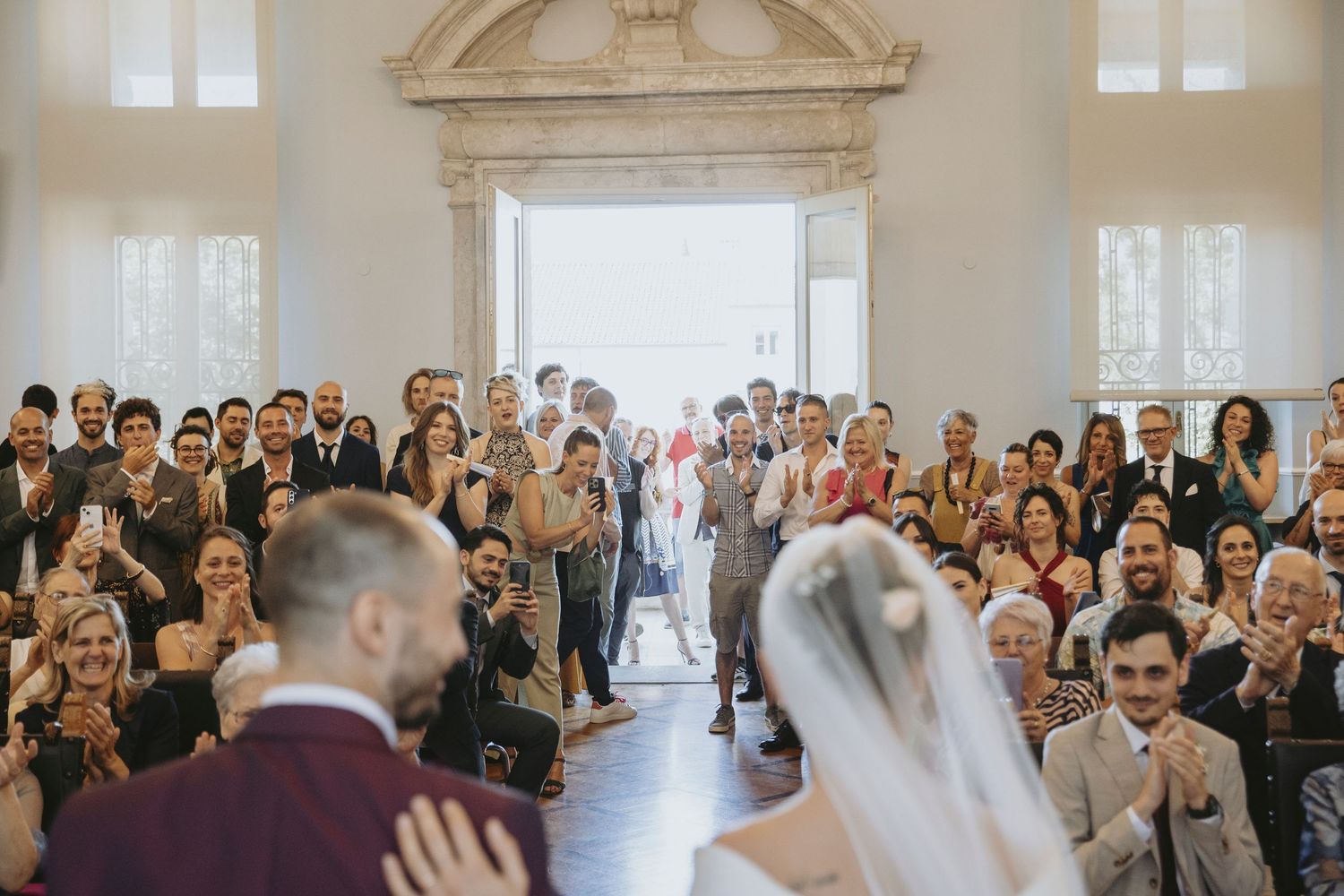 Wedding ceremony guests stand and watch as bride and groom share their first kiss in an elegant white hall.