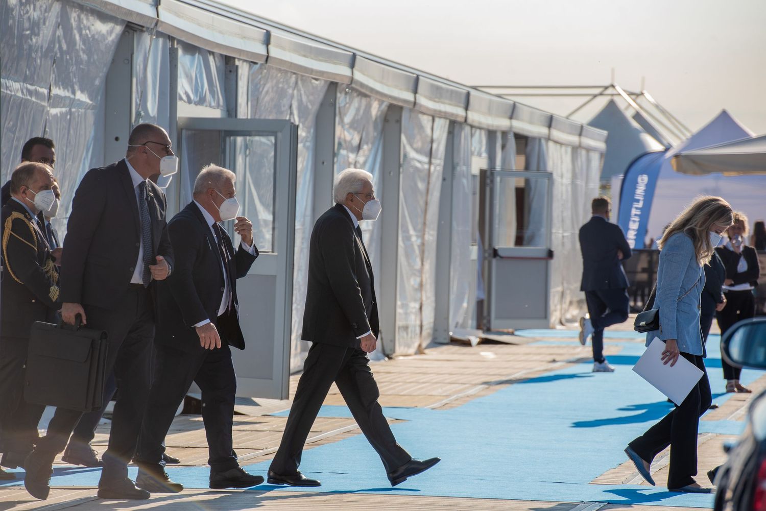 Group of officials in suits and face masks walking along a blue carpet between white event tents outdoors.