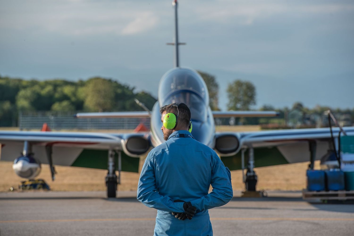 Airport ground crew member in blue uniform and green ear protection stands before a large commercial aircraft on the tarmac.