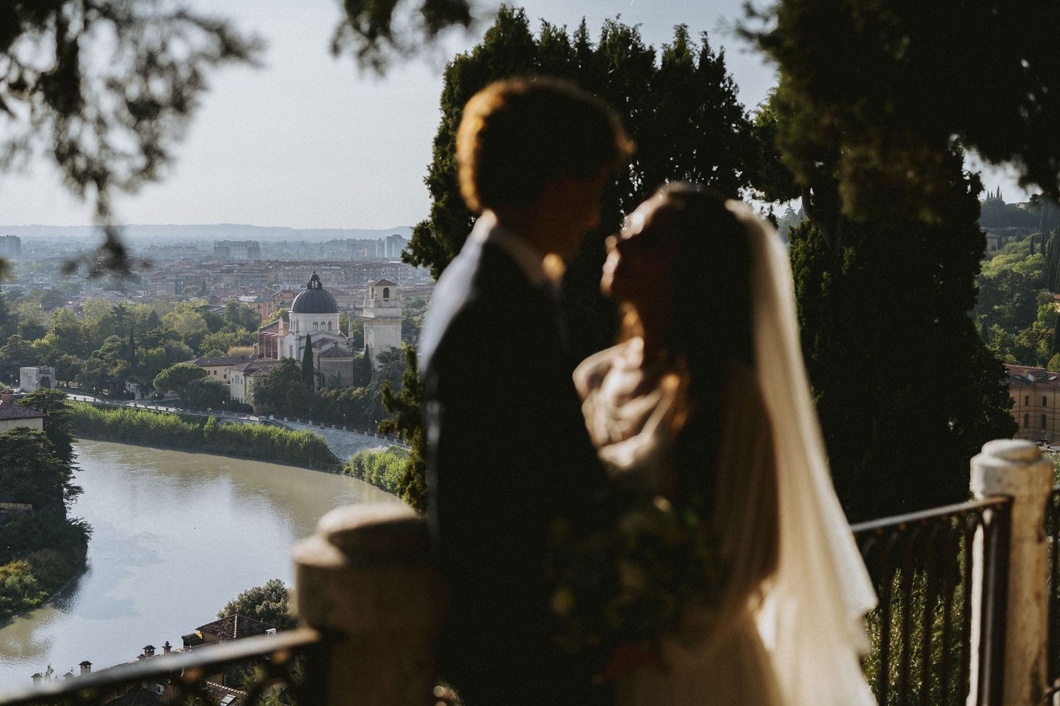 Romantic wedding couple sharing a tender moment with a scenic Italian cityscape and river in the background.