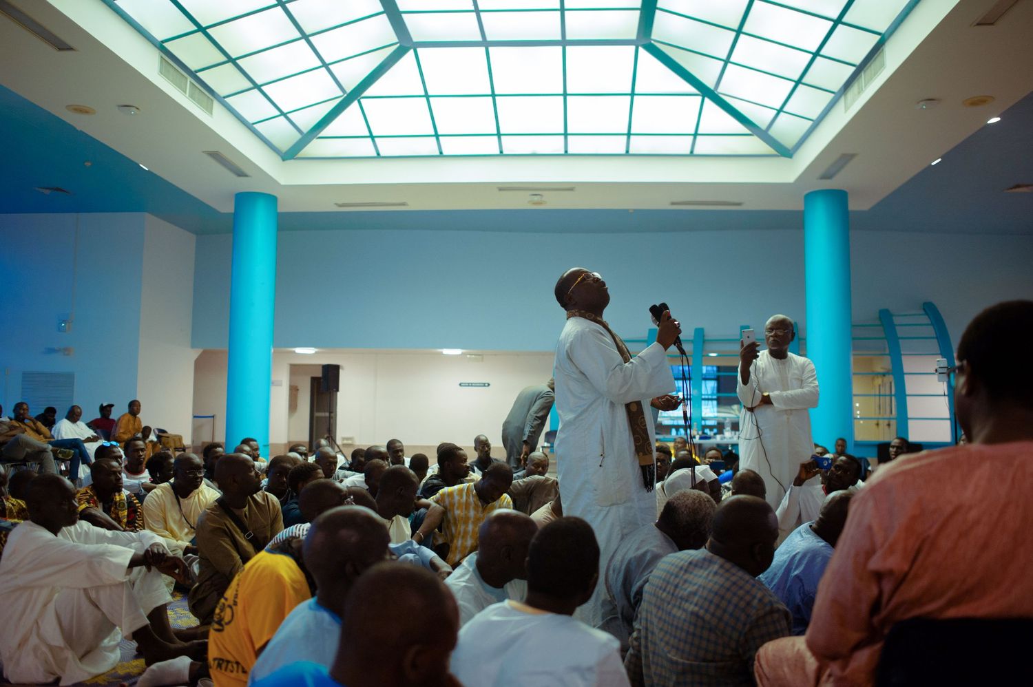 Crowded indoor gathering with speaker addressing large audience in modern hall with blue columns and skylight.